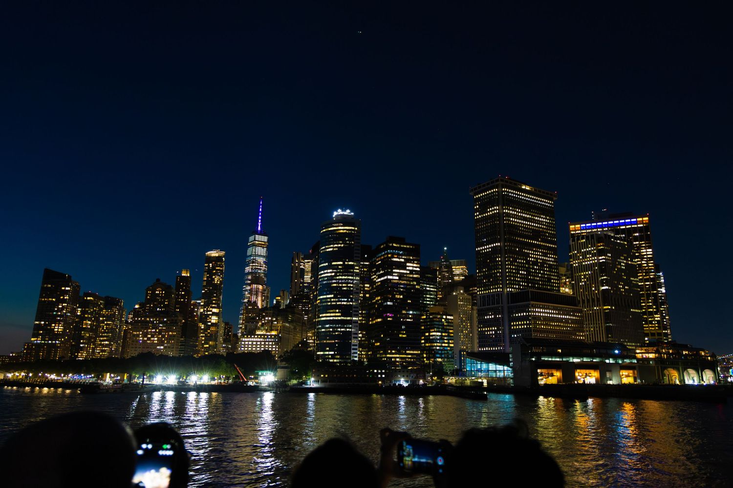NYC skyline glowing at night with illuminated skyscrapers reflecting on the water under a deep blue sky.