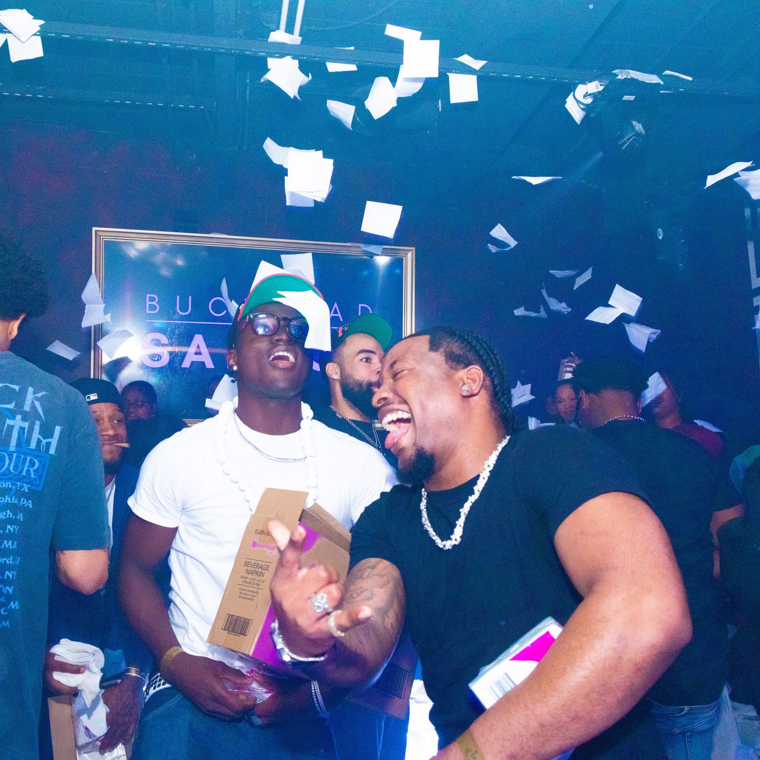 Two men laugh and celebrate at a lively nightclub party with confetti flying in the air.
