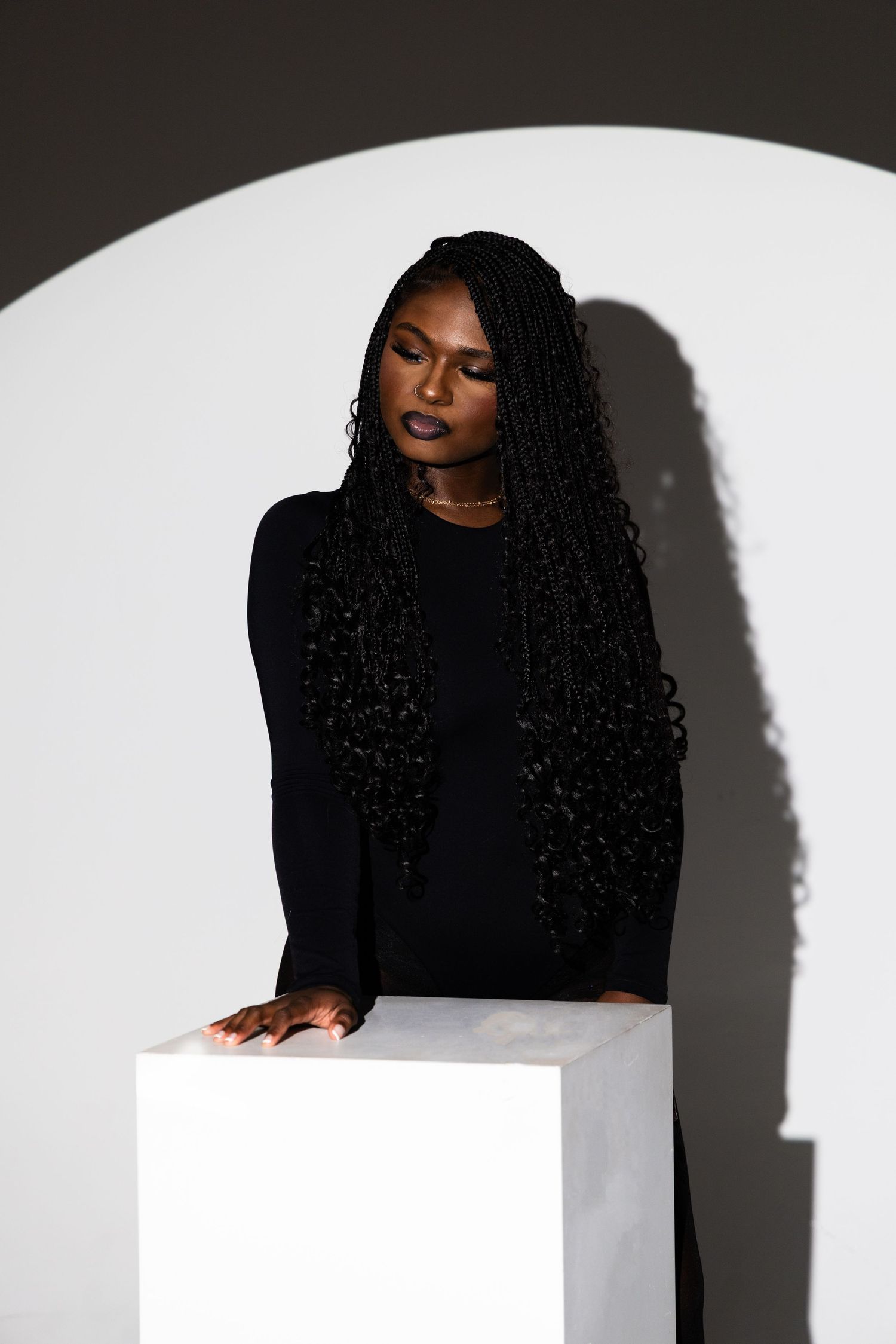 Black woman with long braids in all-black outfit poses elegantly beside a white pedestal against a studio backdrop.