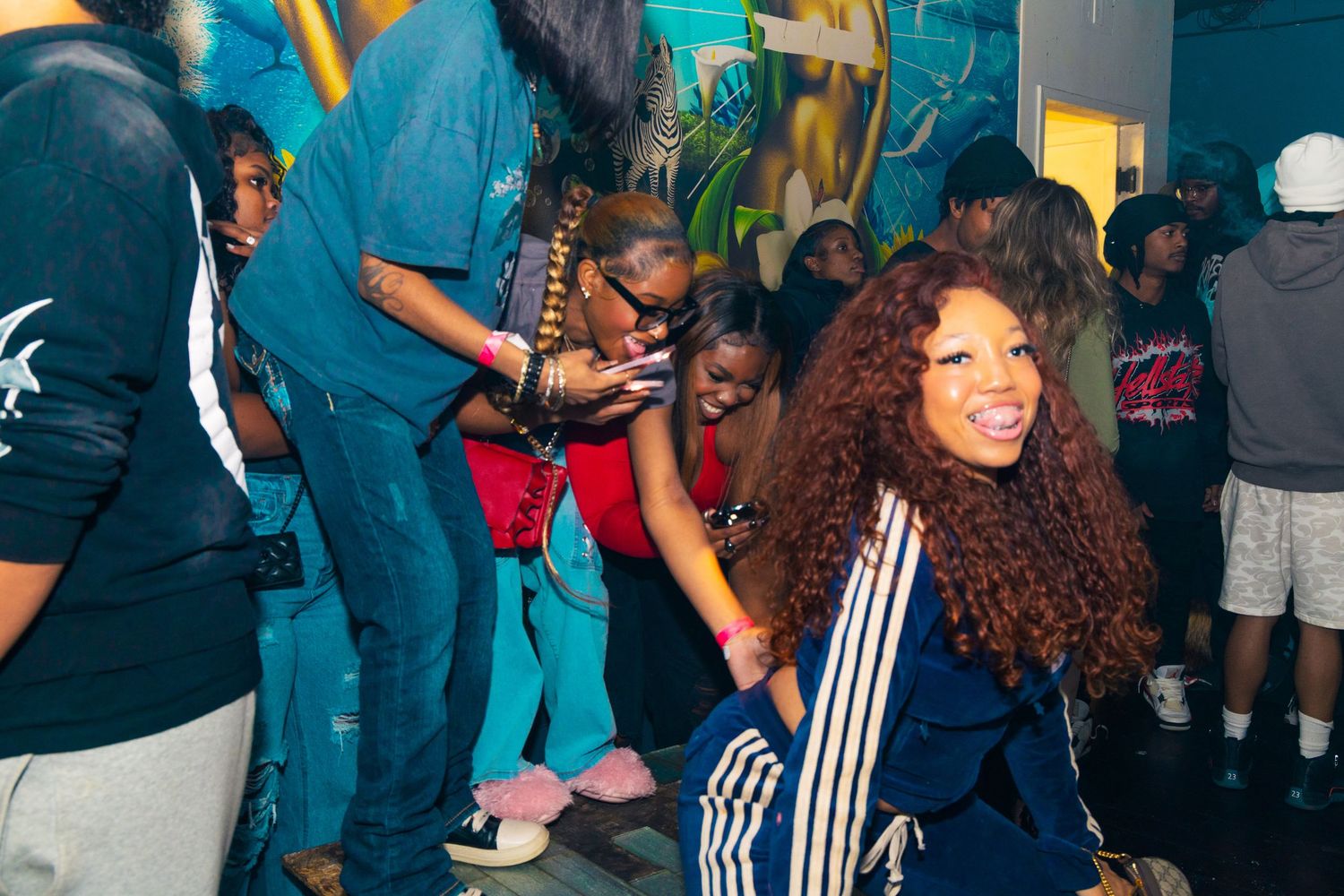 Young woman in blue Adidas tracksuit dancing and smiling at a lively indoor party with friends.