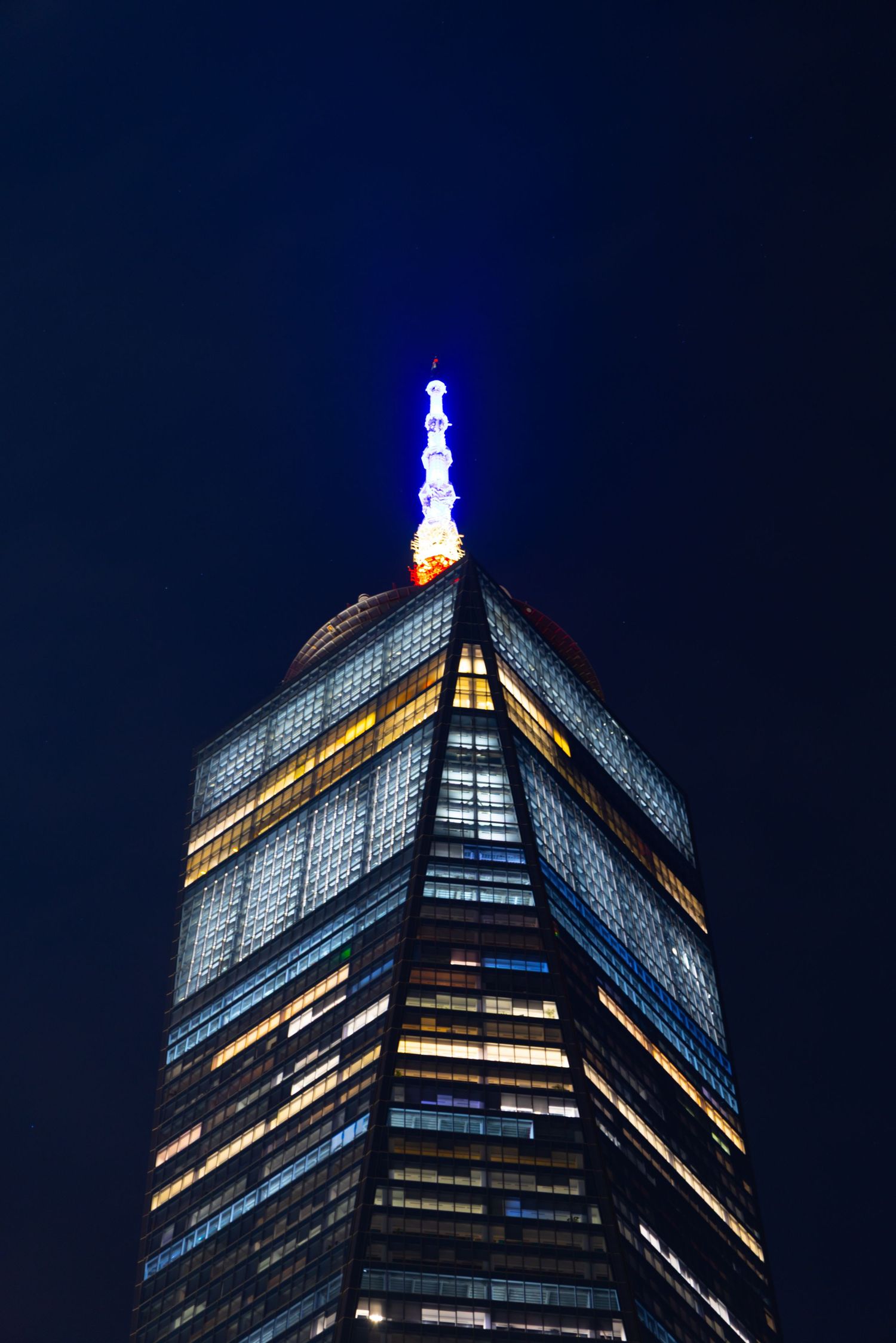Illuminated skyscraper top with glowing blue antenna spire against dark night sky, modern glass tower with lit windows.
