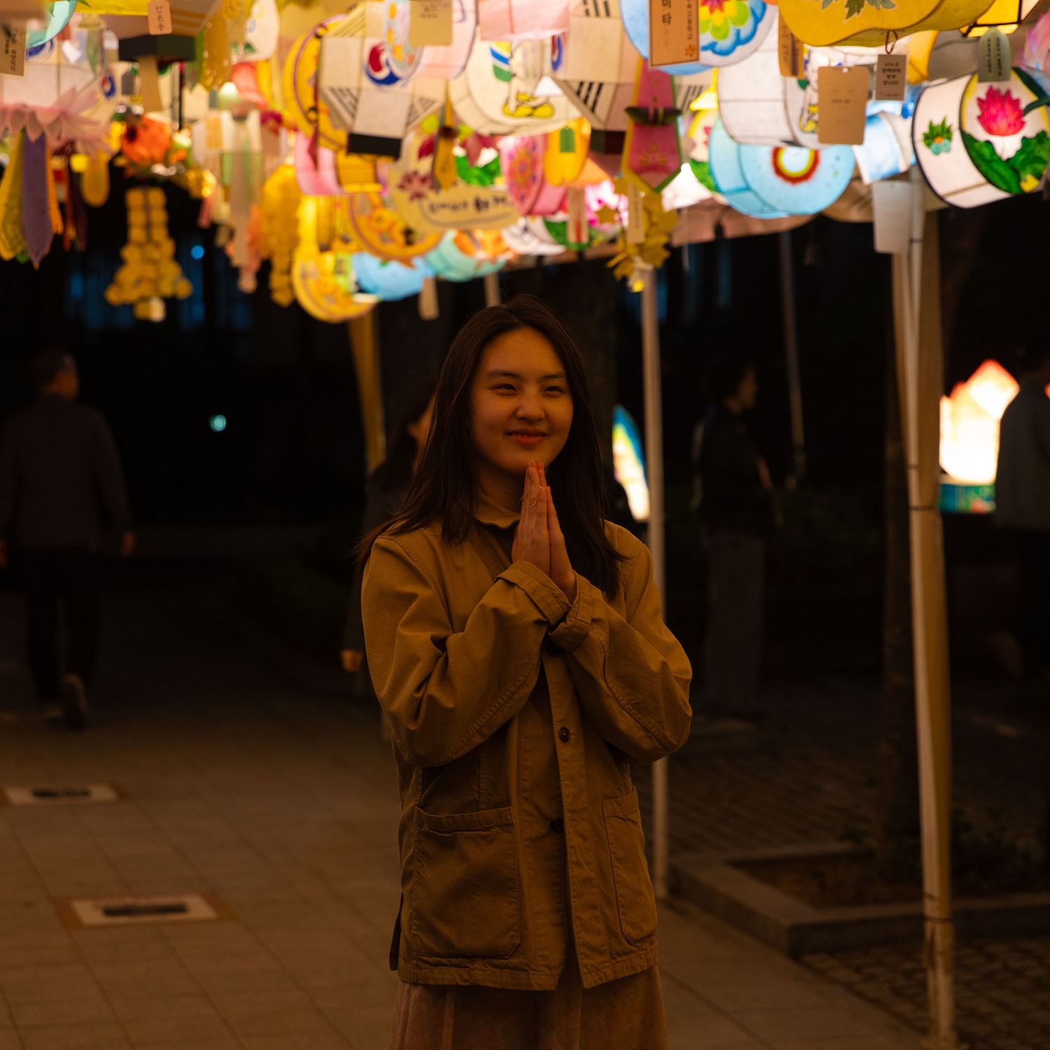 A young woman smiles with hands pressed together in prayer pose under colorful hanging lanterns at night.