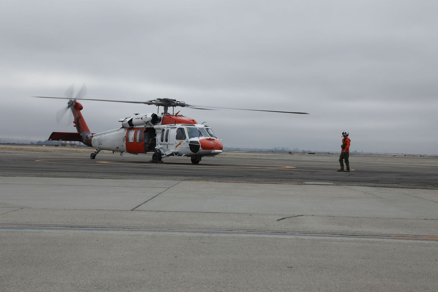 US Coast Guard orange and white Sikorsky MH-60 Jayhawk helicopter on tarmac with ground crew member nearby.