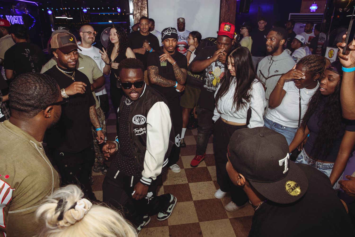 Lively nightclub scene with people dancing and socializing on a checkered floor under dim lighting.