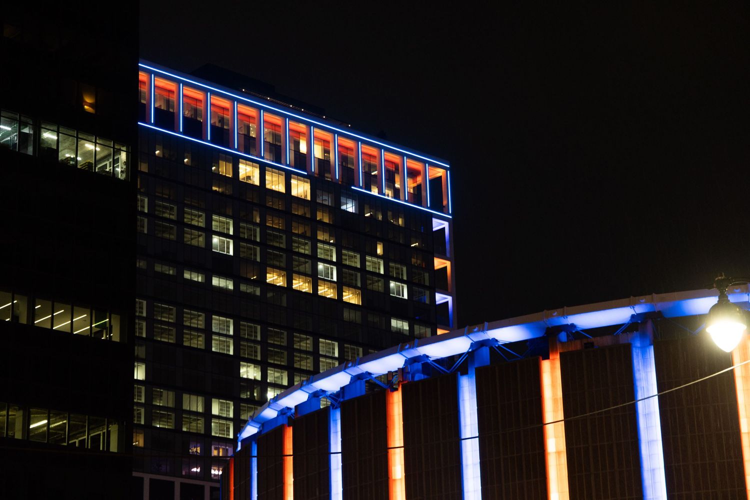 A tall office building illuminated with blue and orange neon lights at night, with glowing columns in the foreground.