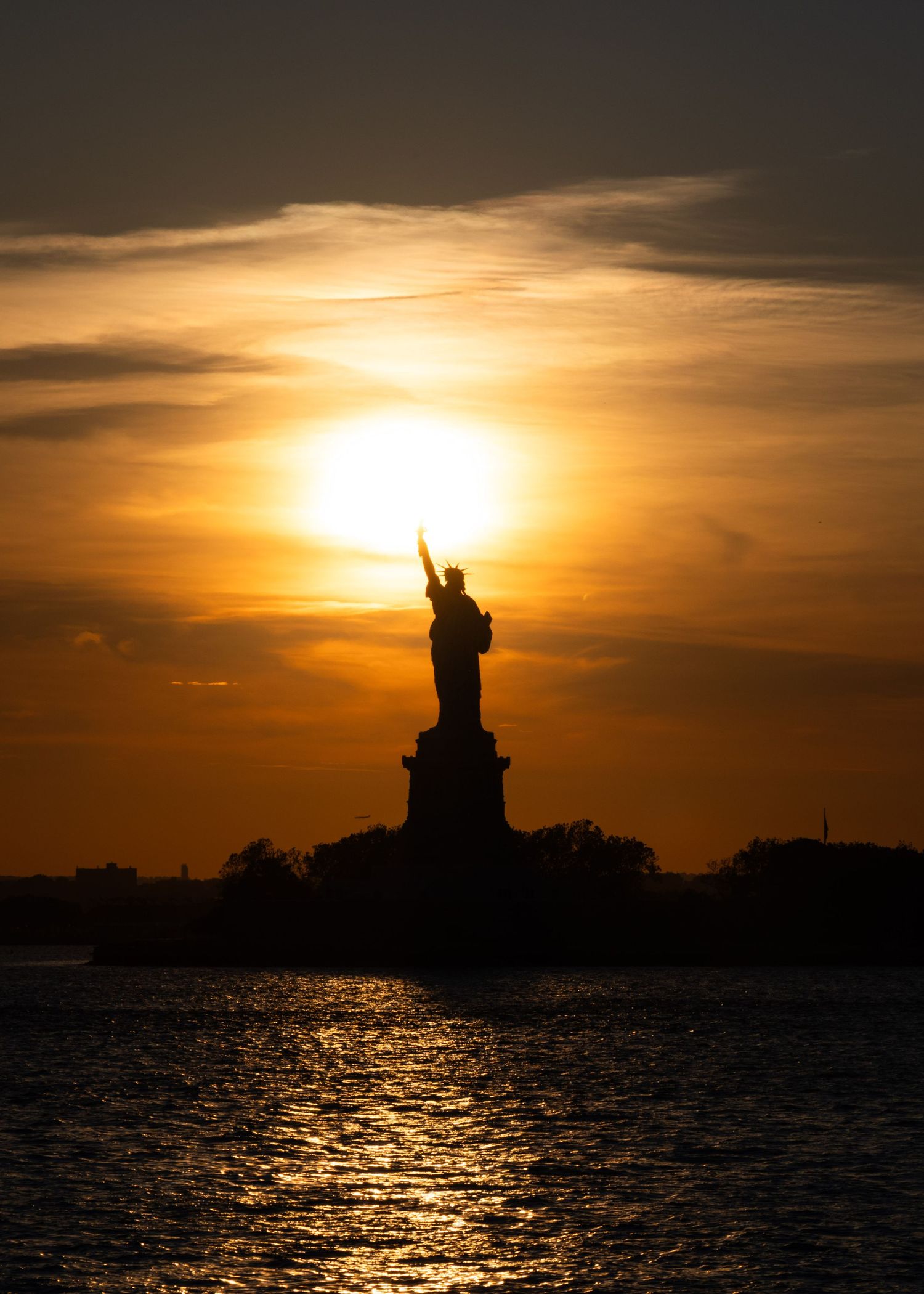 Silhouette of the Statue of Liberty against a golden sunset sky reflected on calm harbor waters.