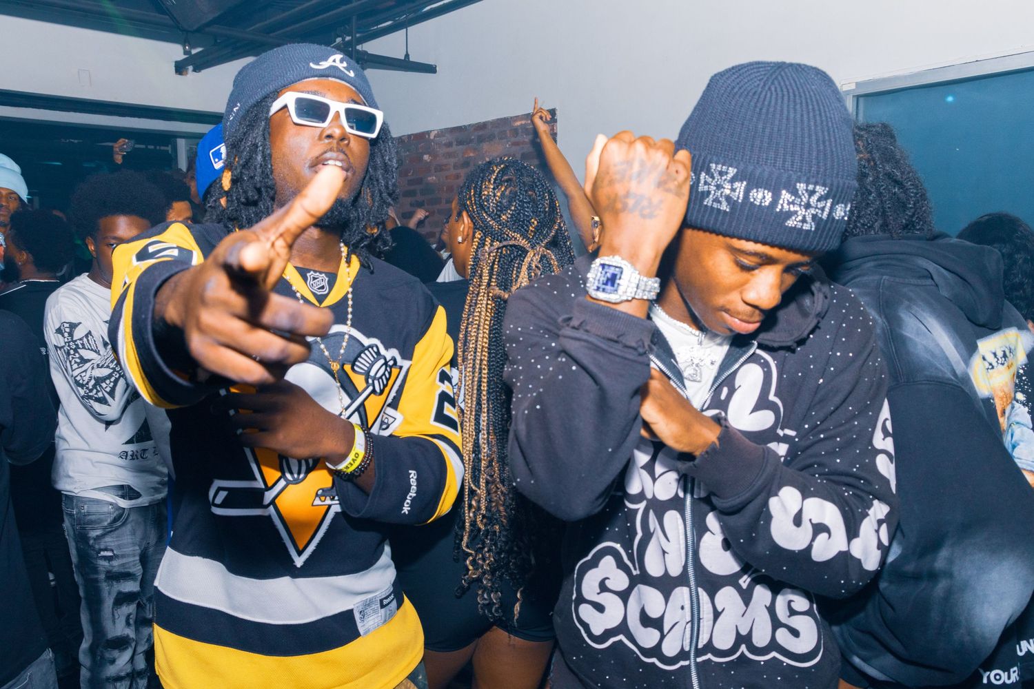 Two young men posing at a party, wearing streetwear including jerseys and hoodies under colorful club lighting.