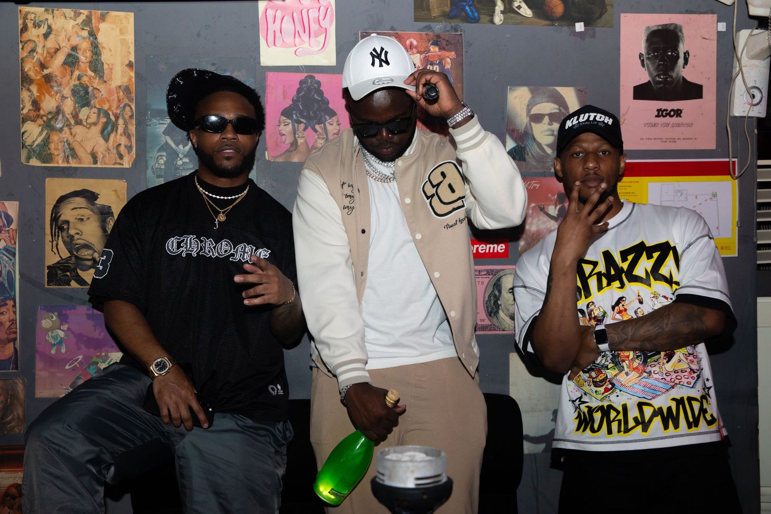 Three men posing with a bottle in front of music posters at a party.