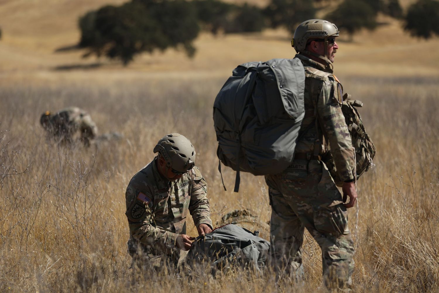 Two soldiers in camouflage gear gather equipment in a dry grass field after a military parachute operation.