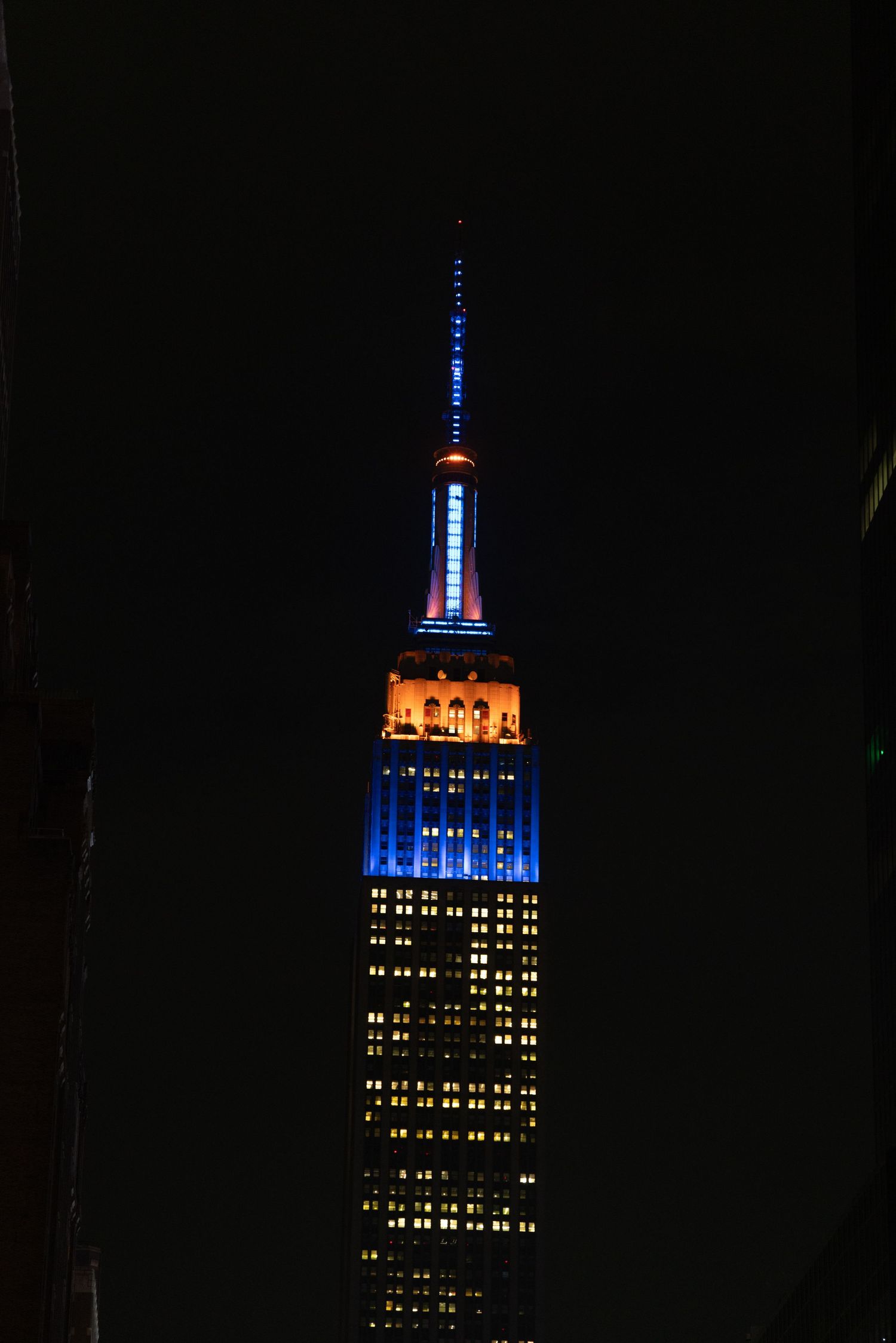 Empire State Building illuminated at night with blue and orange lights against a dark NYC sky.