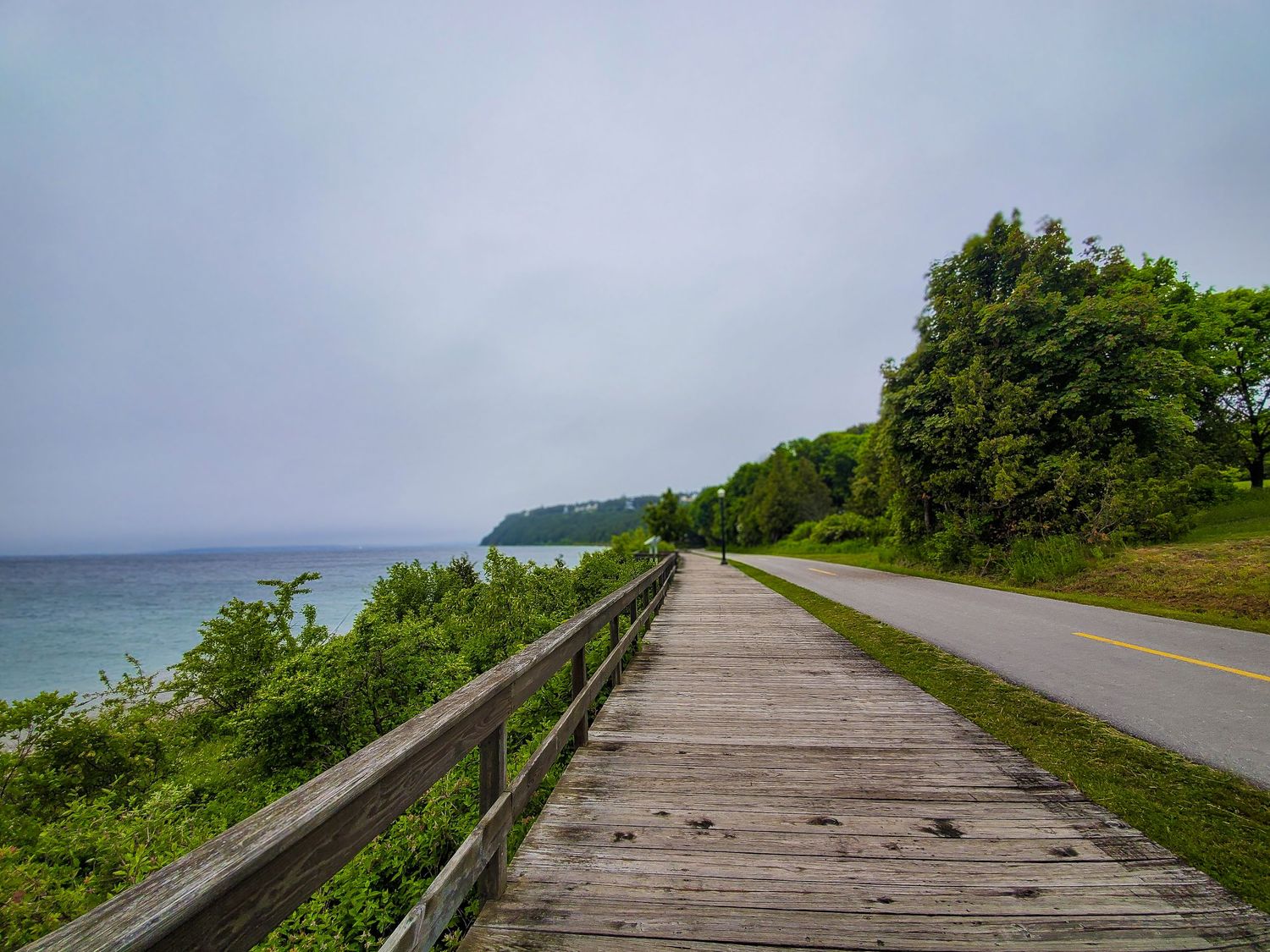A wooden boardw a coastal roa Mackinaw Island, stretches into the distance with ocean views and lush greenery on both sides.