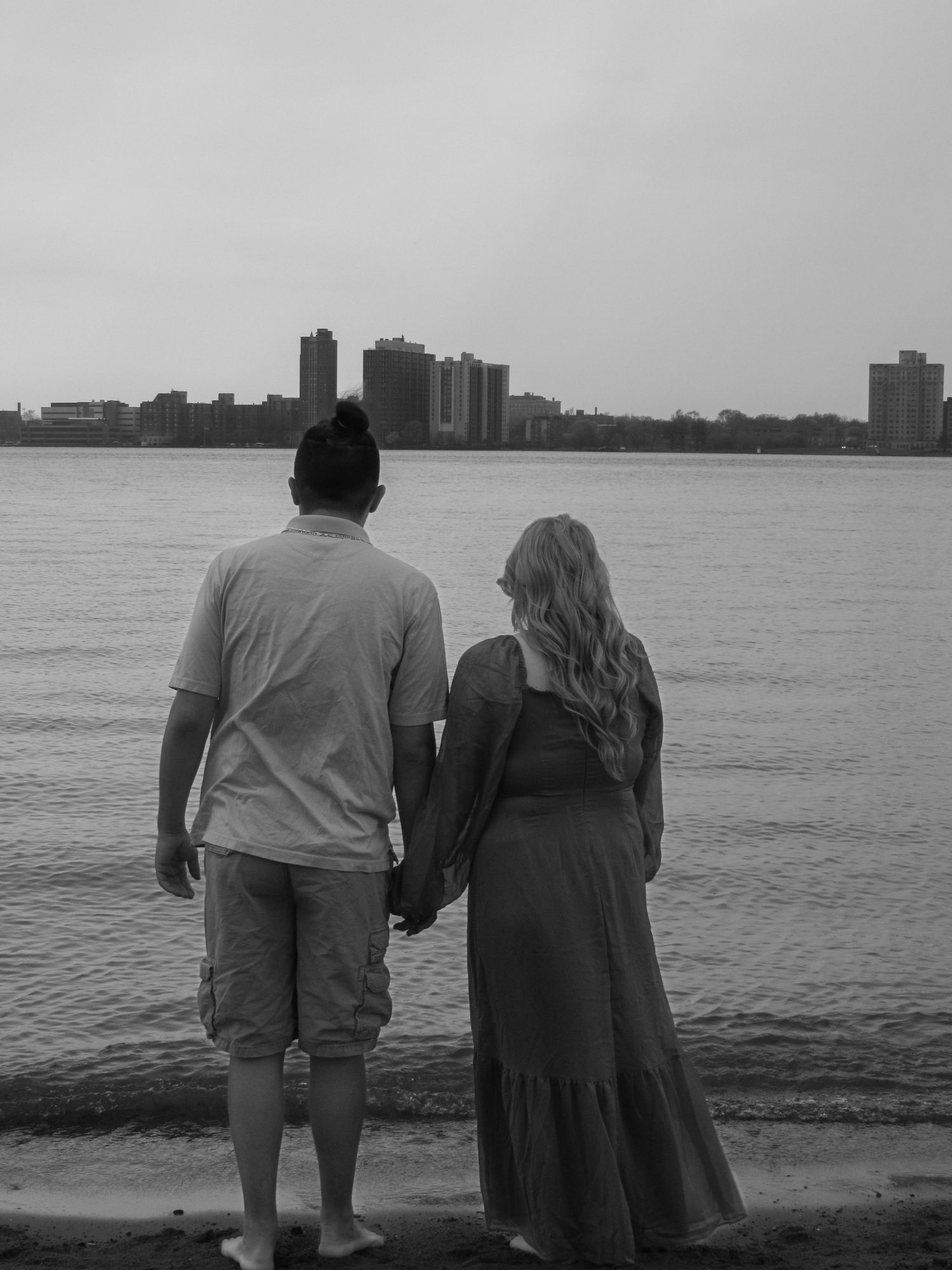A couple stands at the edge of a waterfront, holding hands while gazing at the distant city skyline across the water.