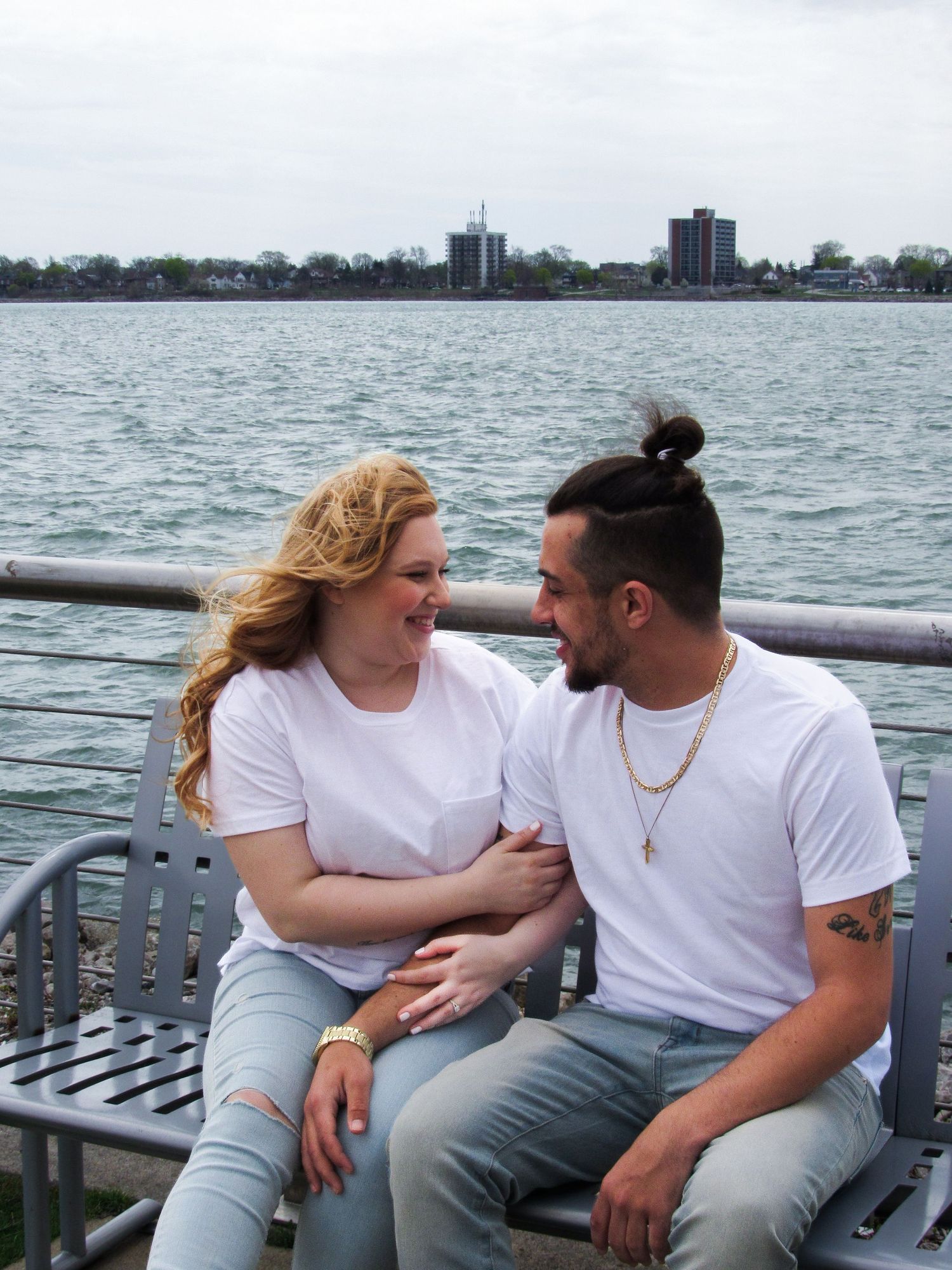 A couple sits together on a waterfront bench with a city skyline visible in the distance.