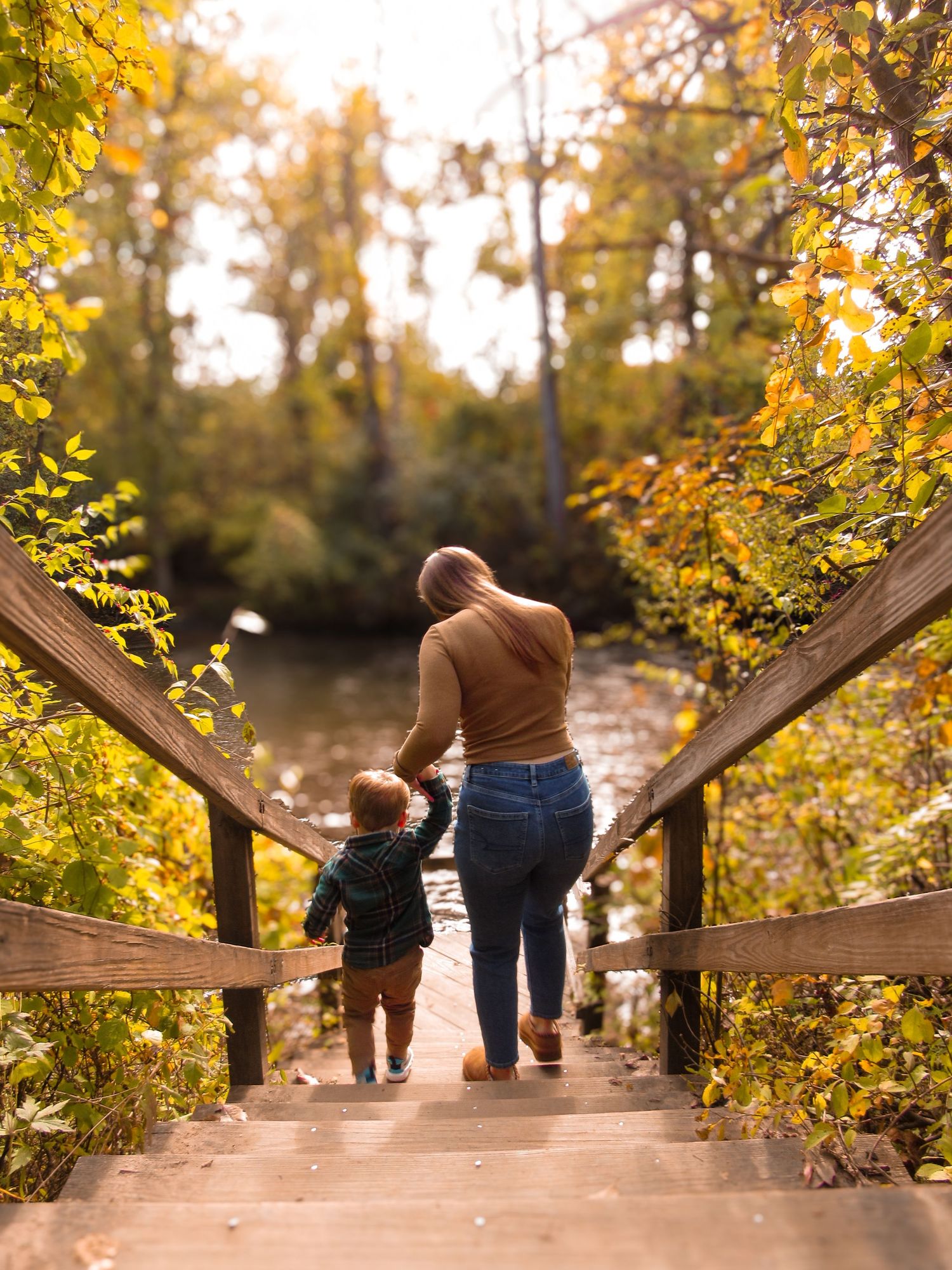 Mother & son walking down wooden steps surrounded by golden fall trees near a river.