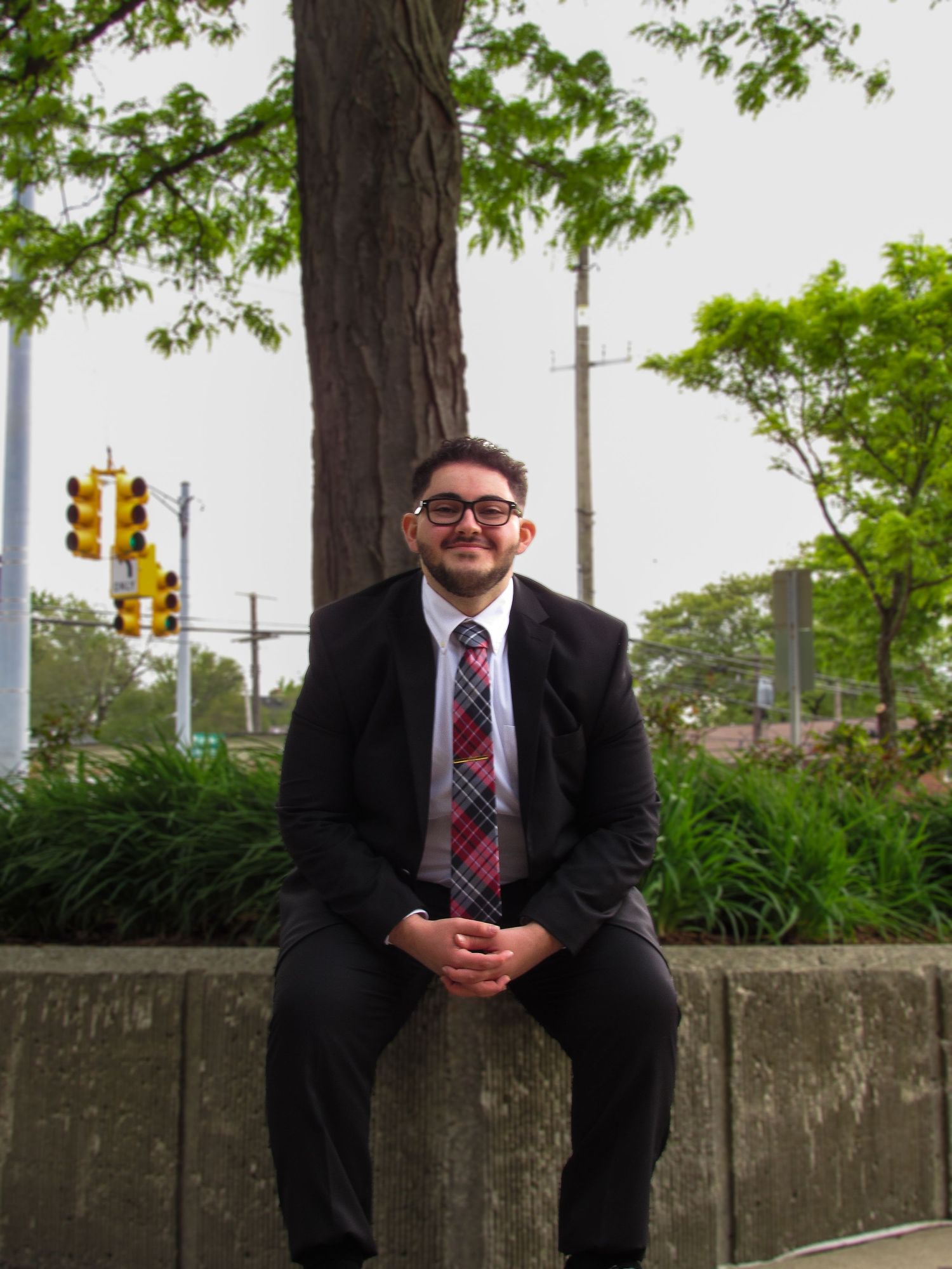 Professional in a dark suit and red tie sitting on a concrete wall outdoors near trees.