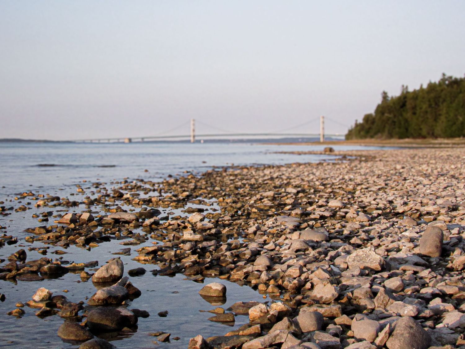 Rocky shoreline along lake Michigan with the Mackinaw bridge visible in the distance under a pale sky.