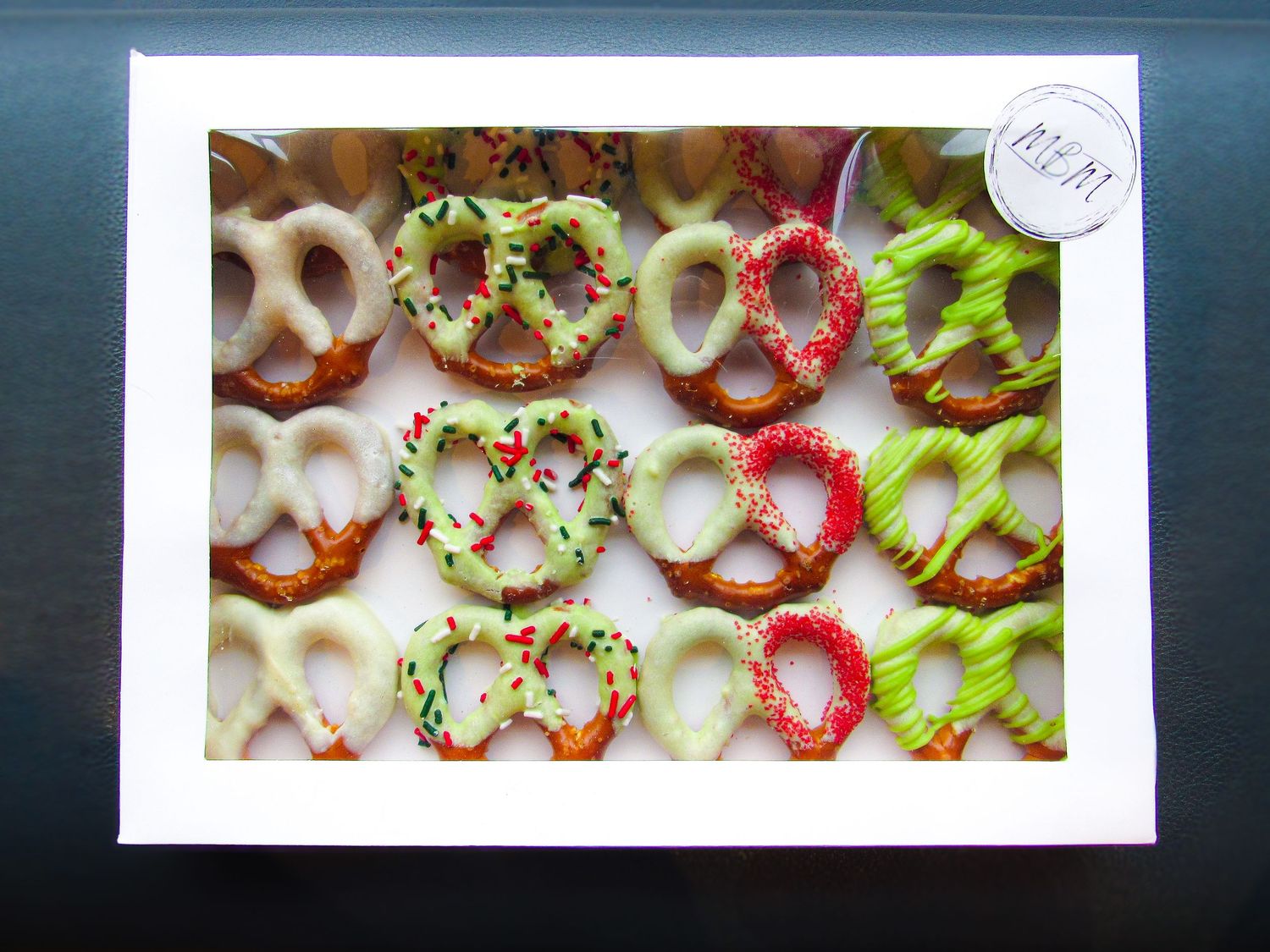 Chocolate covered pretzels decorated with colorful sprinkles and toppings arranged neatly in a white box.