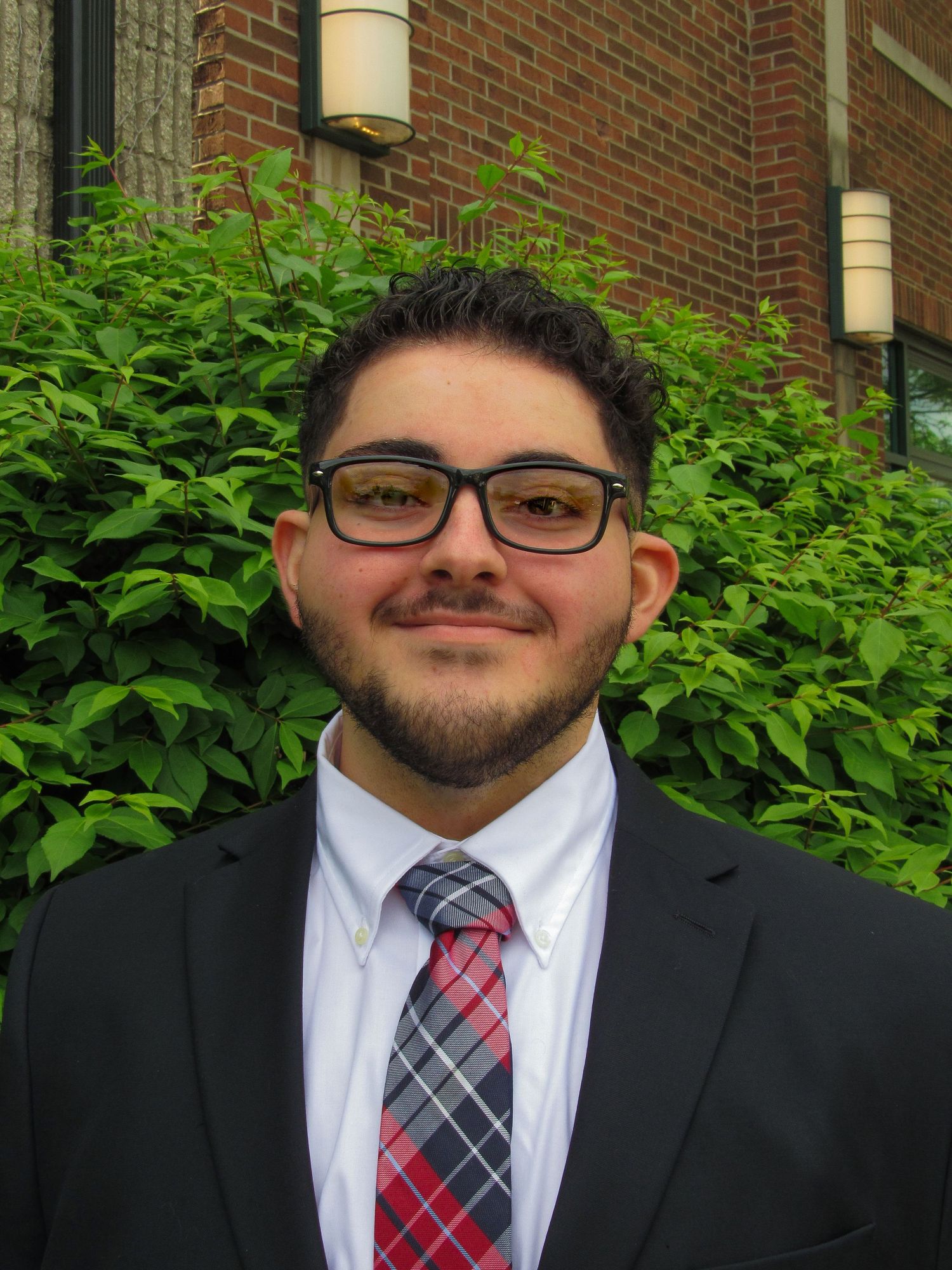 Professional wearing a black suit with striped tie poses against brick wall with green foliage.