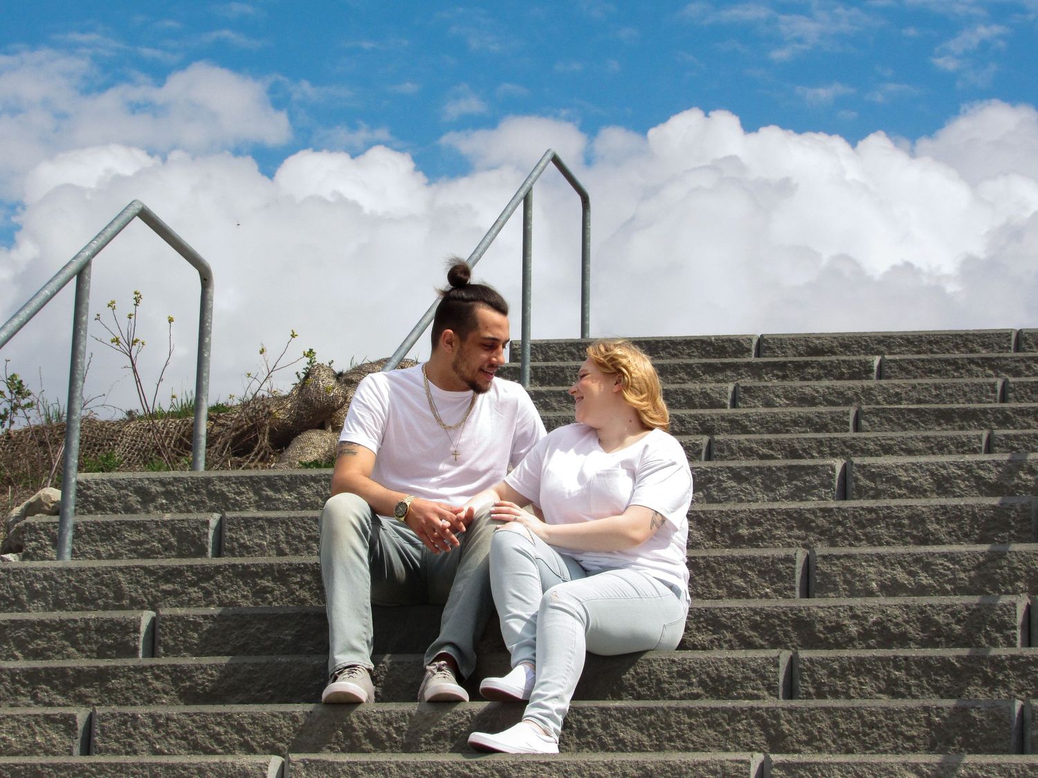 Two people share an intimate moment on concrete steps against a blue sky.