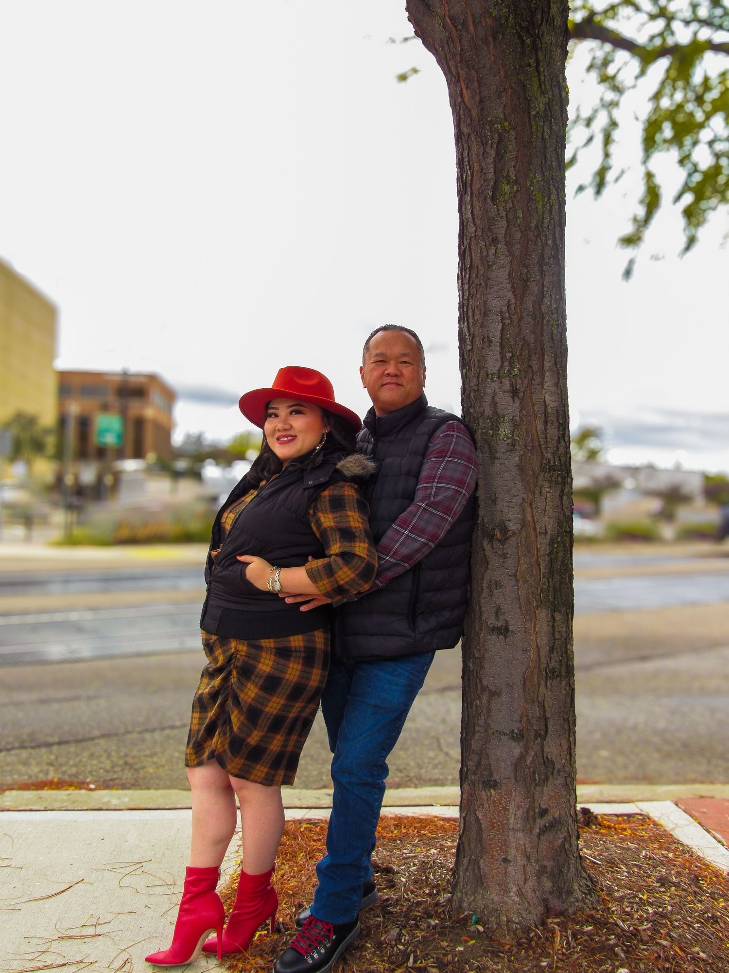 A couple in casual attire share an intimate moment against a tree trunk in an outdoor setting.
