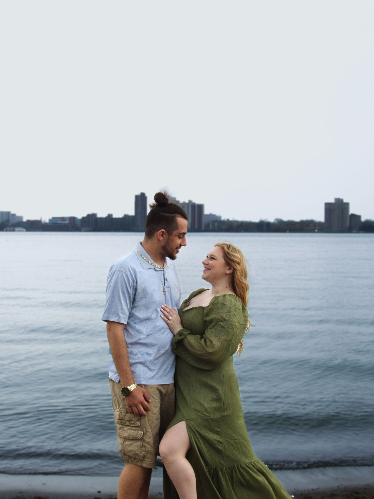 A couple poses romantically by a waterfront with a cityscape in the background at dusk.