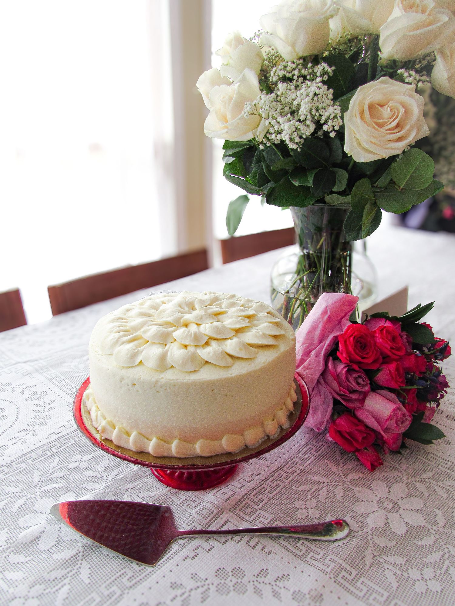 White frosted cake with delicate piped decorations sits on a red plate next to pink roses on a lace tablecloth.