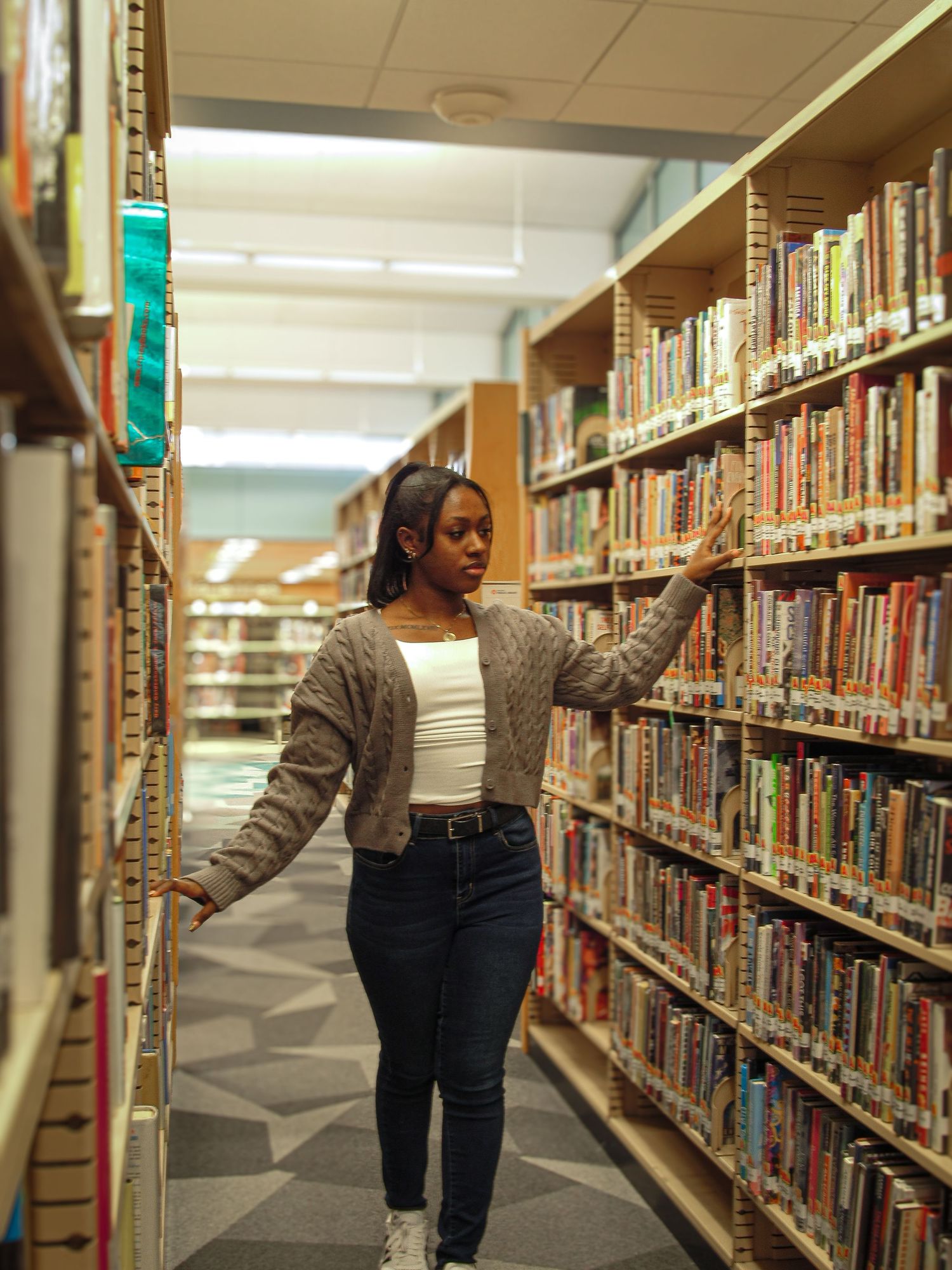 Person browsing books in a library aisle with fluorescent lighting overhead.