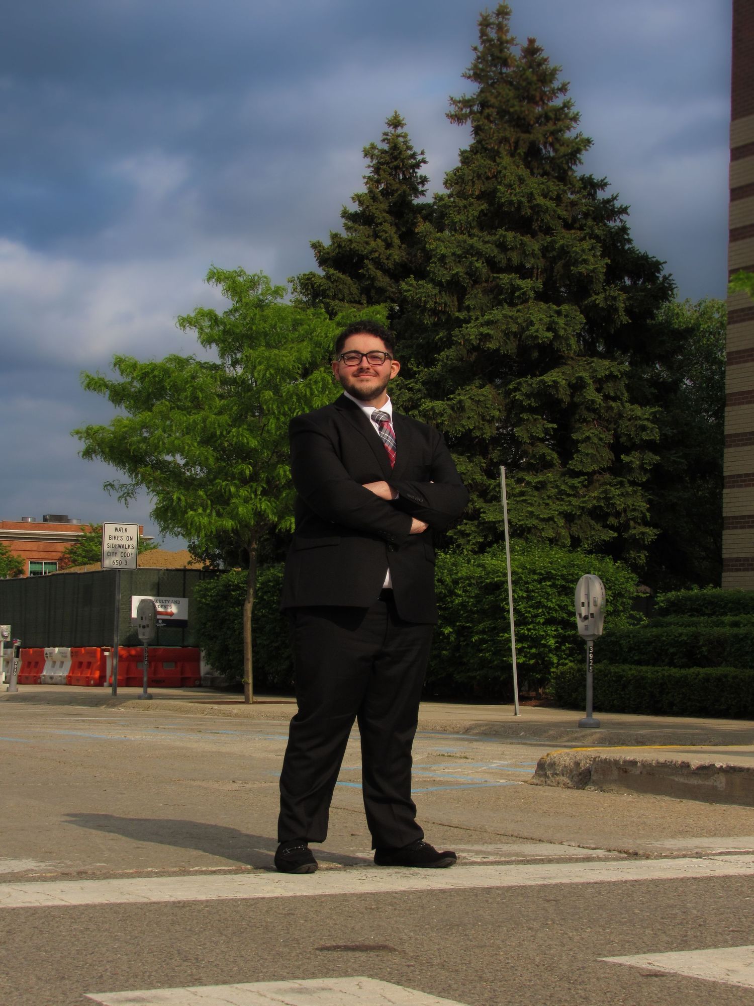 A figure in a dark business suit stands in an empty street with evergreen trees and buildings in the background.