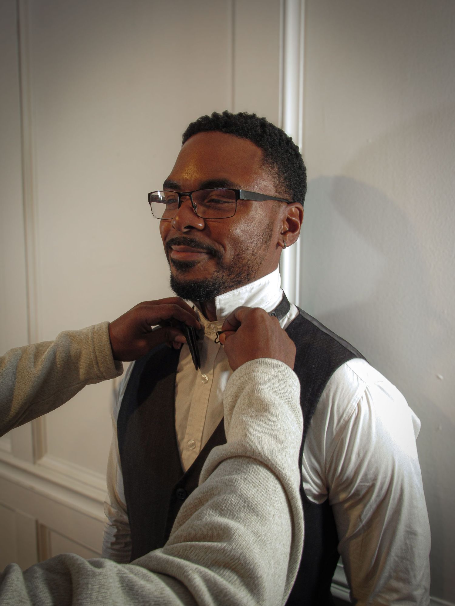 Someone adjusts a bowtie while wearing glasses, a white dress shirt and dark vest in an indoor setting.
