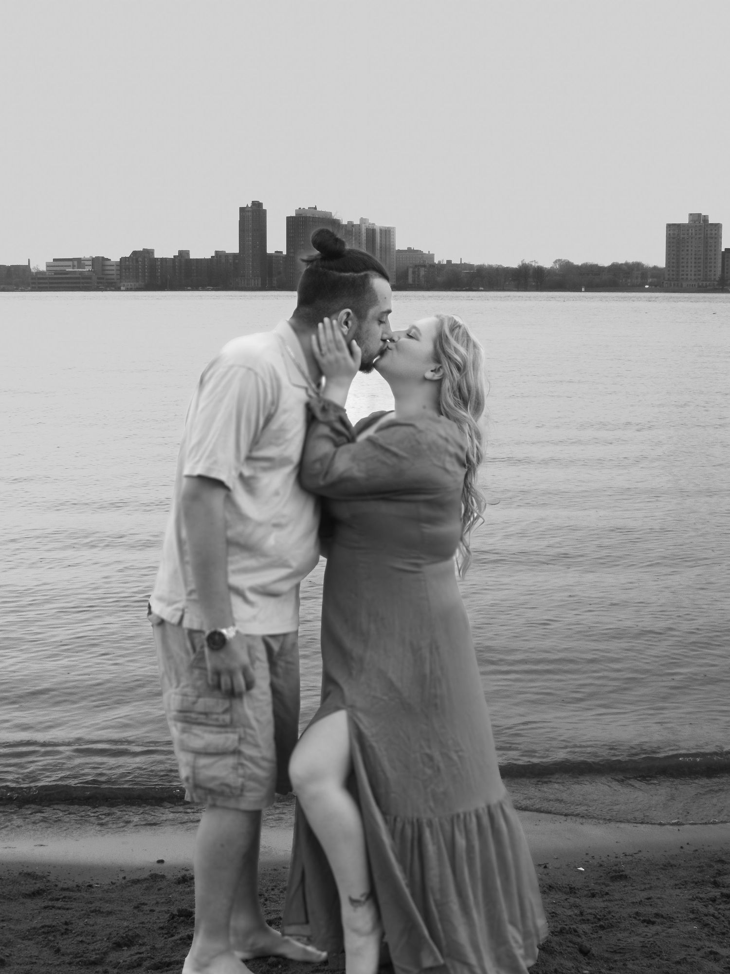A couple embraces on a beach with a city skyline visible in the distance during sunset.