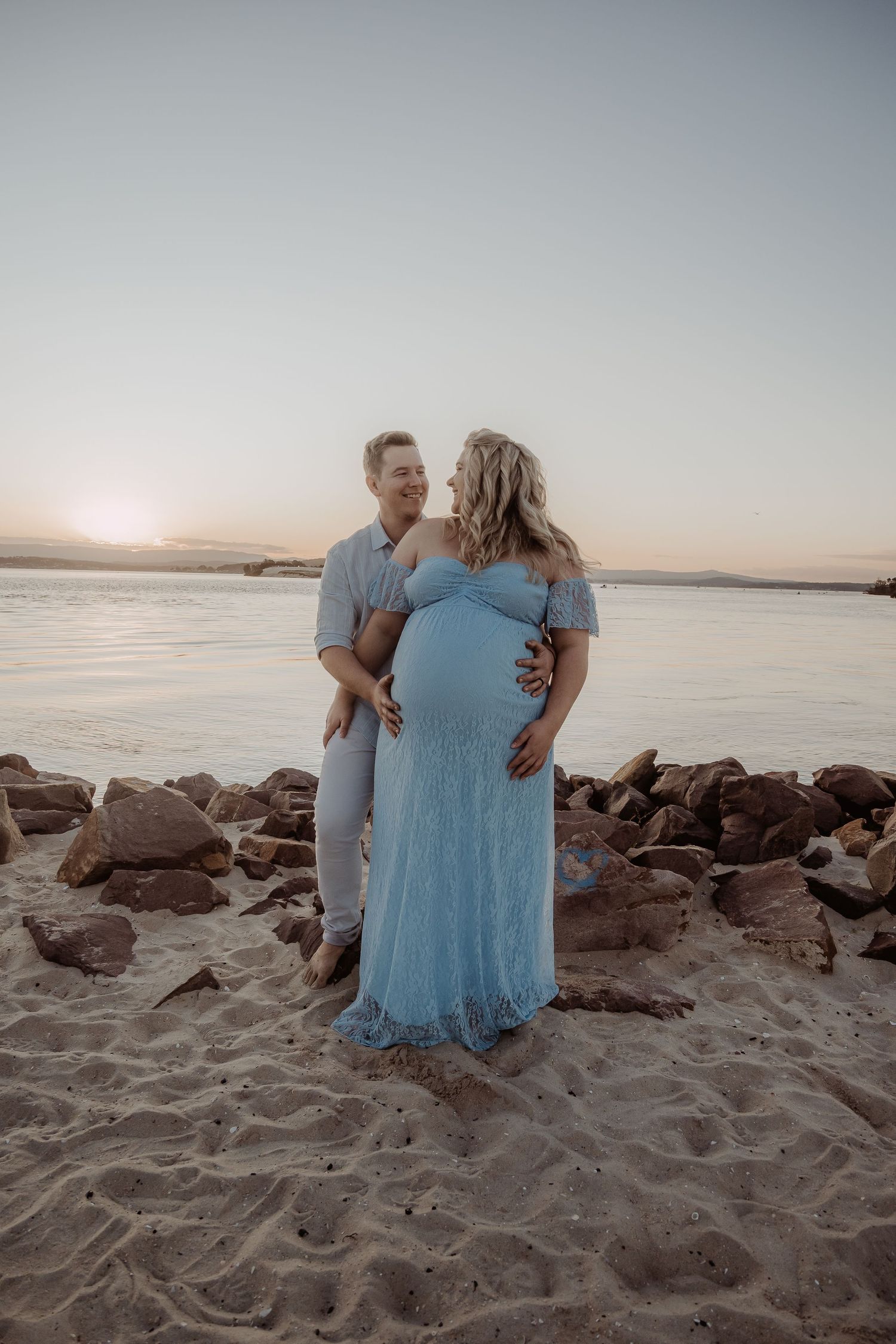 Couple embraces on a beach at sunset with rocks and water in background, wearing light blue attire at a newcastle maternity photoshoot.