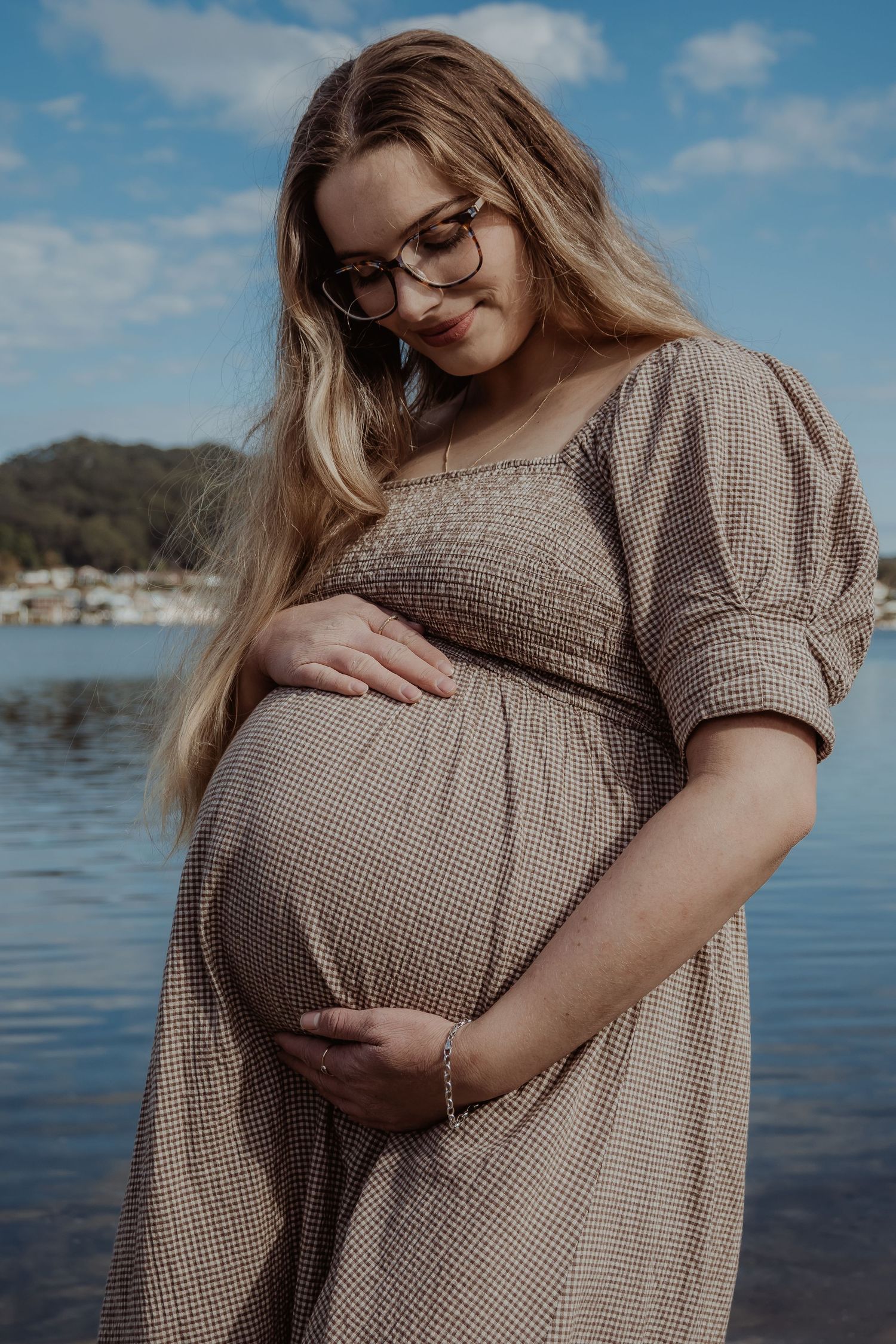 Pregnant woman in checkered dress poses by waterfront on cloudy day during central coast maternity session.