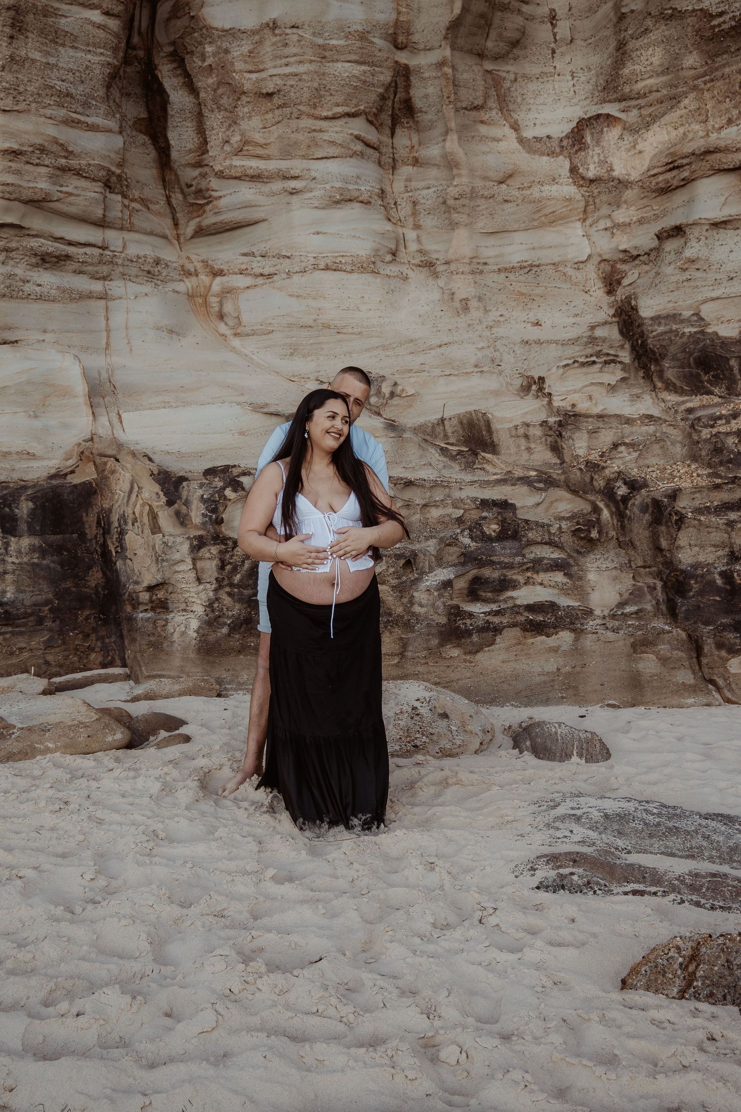 A couple embraces against dramatic rocky cliffs at the beach in a romantic central coast maternity photoshoot series.