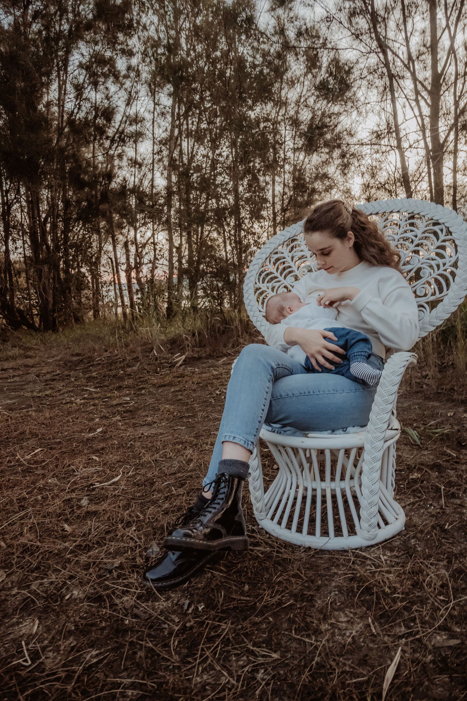 Mum in jeans and a white top poses on a vintage white wicker chair in a wooded outdoor setting at a central coast breastfeeding photoshoot.