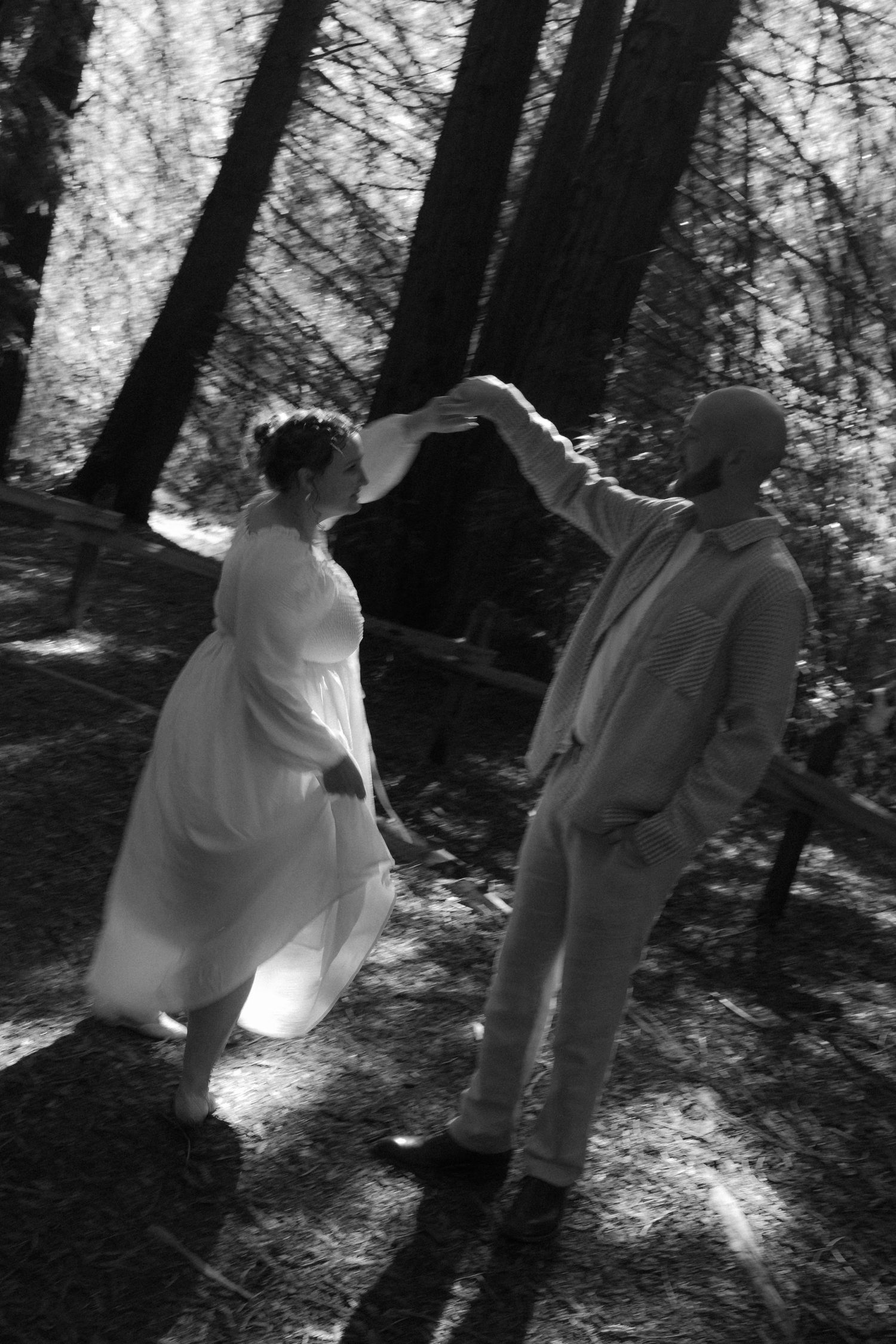 A couple dances together in a sunlit forest clearing in black and white.