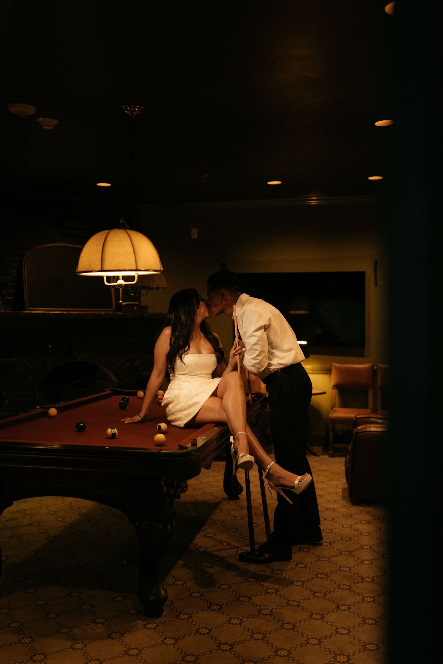 Couple sharing a romantic kiss on a pool table in a dimly lit room with warm vintage lighting.