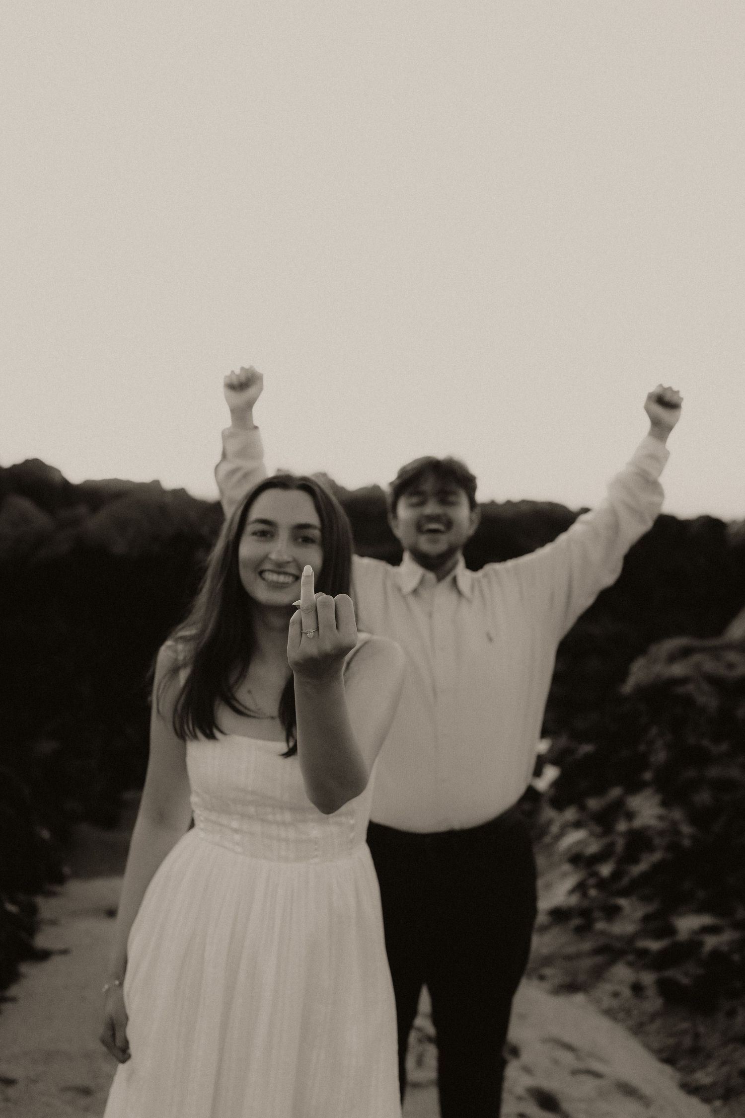 Black and white photo of smiling bride showing ring while groom celebrates with raised fists behind her.