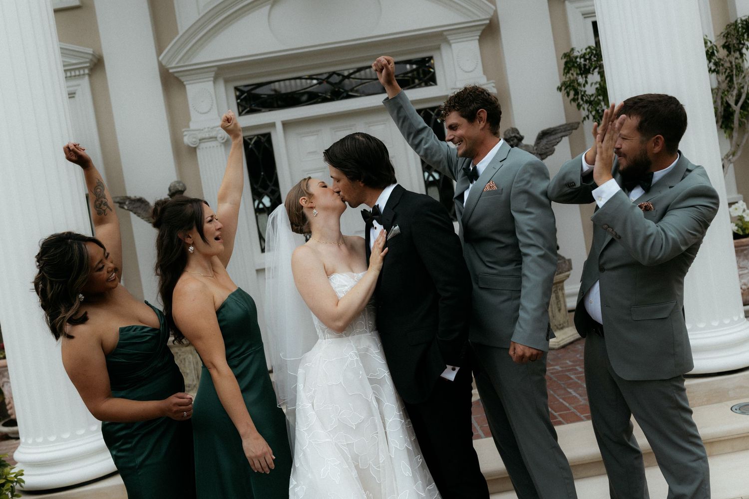 Wedding couple kissing surrounded by cheering bridesmaids in green dresses and groomsmen in gray suits outside elegant venue.