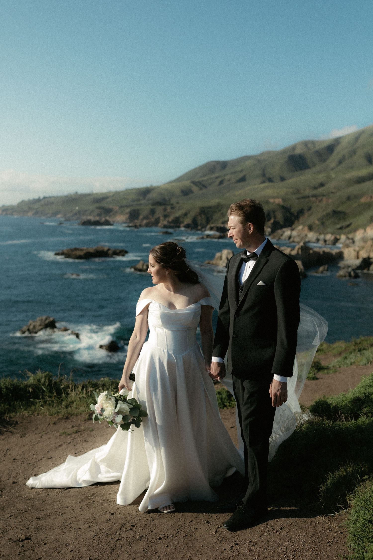 A bride and groom hold hands on a coastal cliff overlooking the Pacific Ocean, with green hills and rocky shores behind them.
