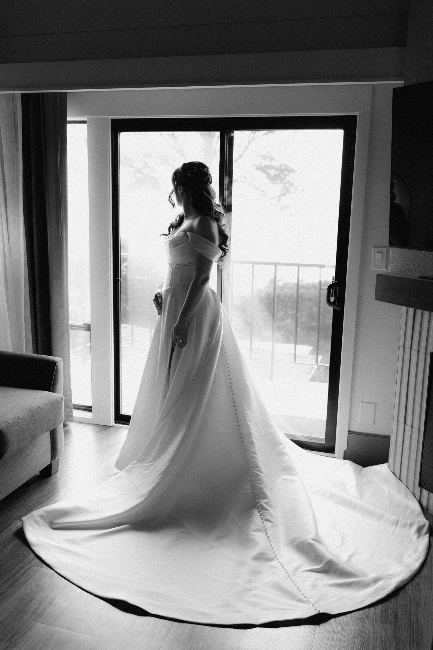 Black and white photo of a bride in a long white gown with train standing by a balcony door.