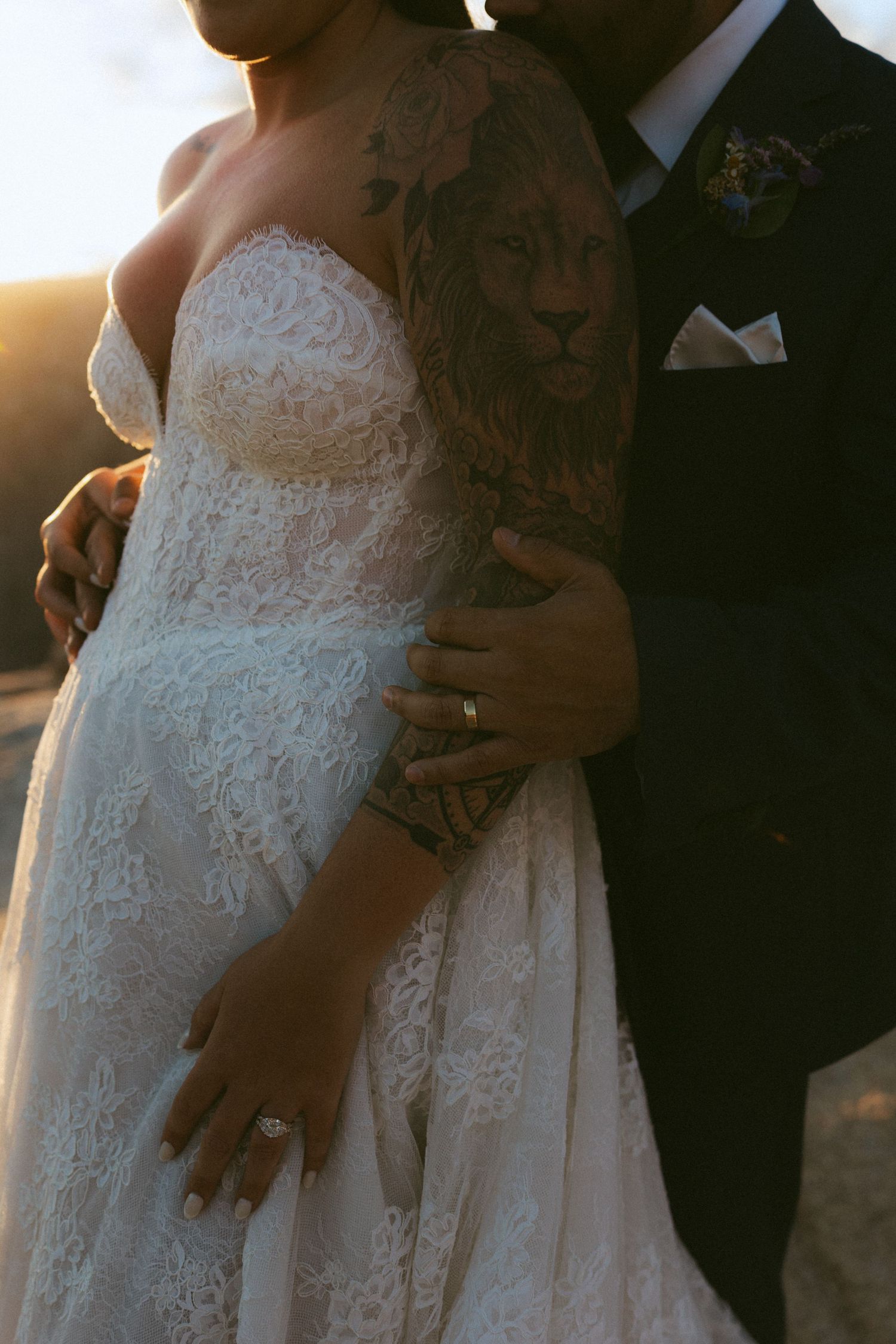 Groom embraces bride in lace wedding dress, showcasing lion arm tattoo at sunset.