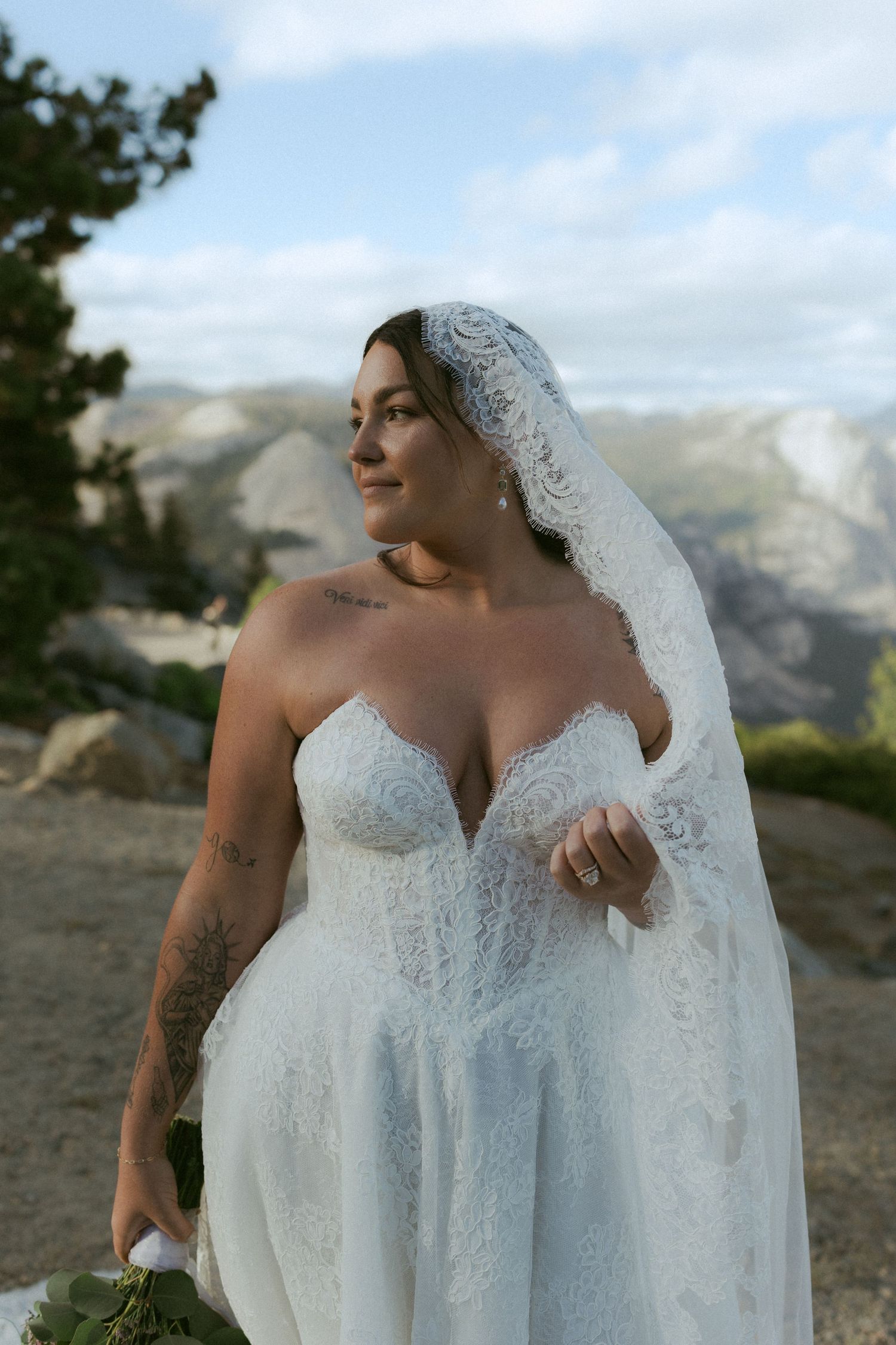 Bride in lace wedding dress and veil poses at Yosemite overlook with mountain views behind her.