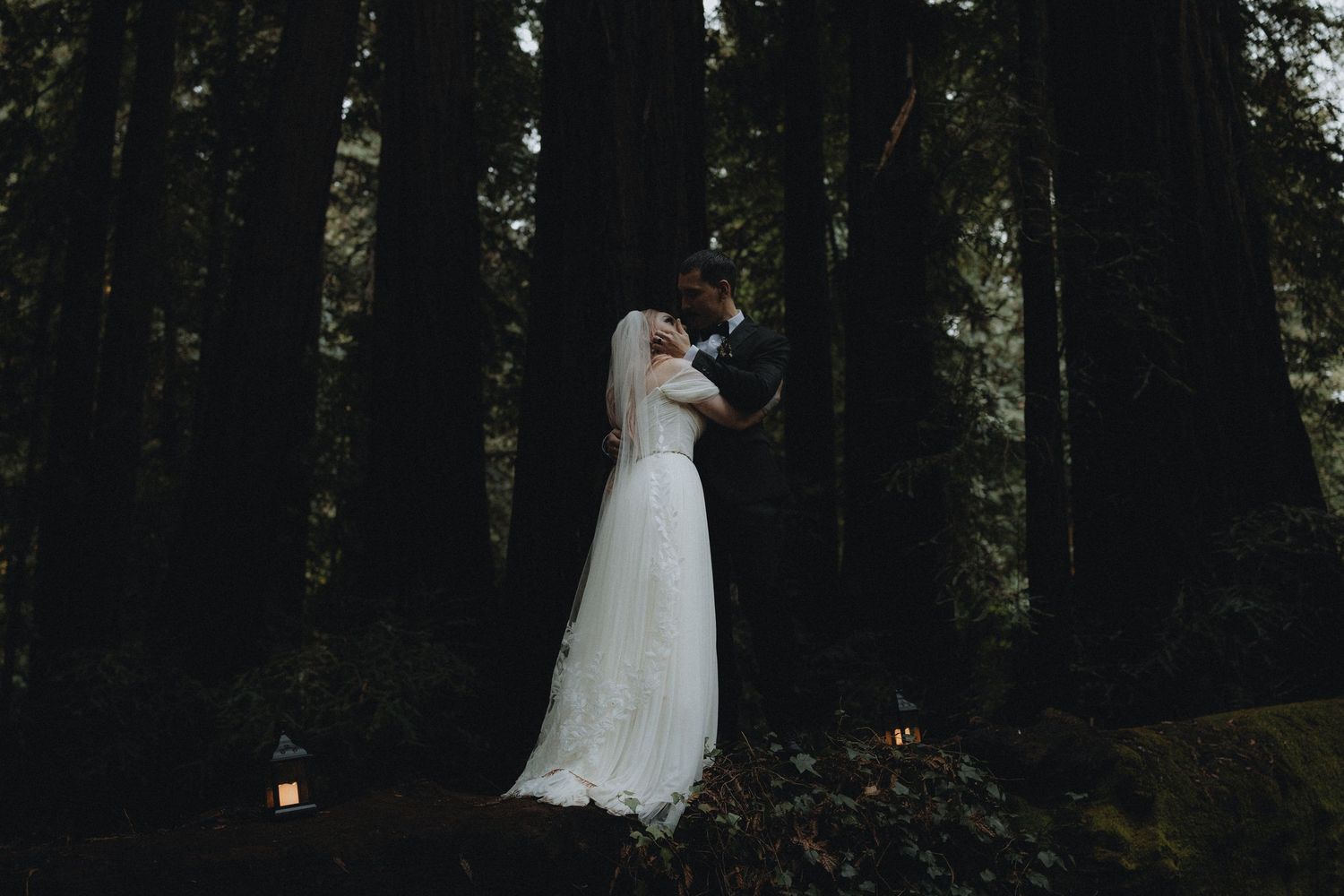 A romantic wedding couple embraces in a moody forest setting with lanterns glowing on the mossy ground.