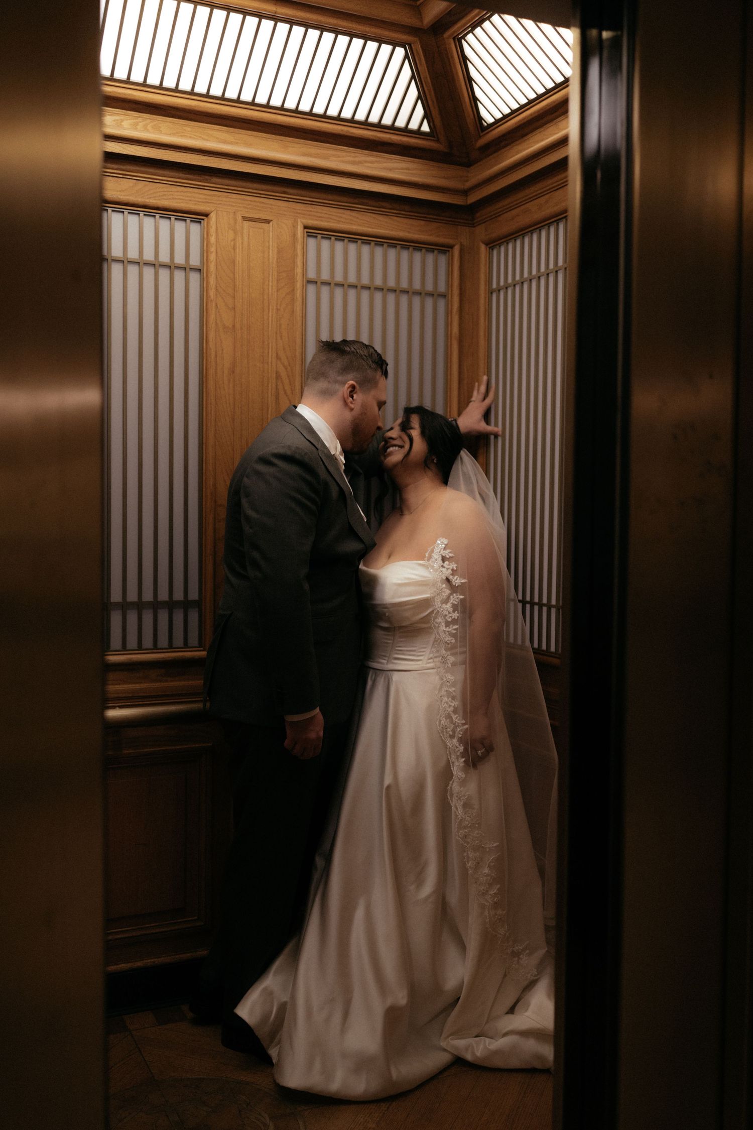A wedding couple shares tender moments in a wood-paneled elevator with vertical lighting.