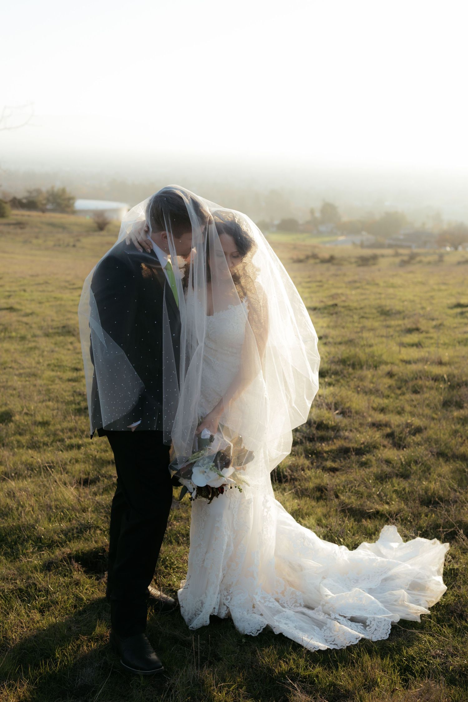 A couple shares an intimate moment under a flowing veil in a misty field during their wedding photoshoot.