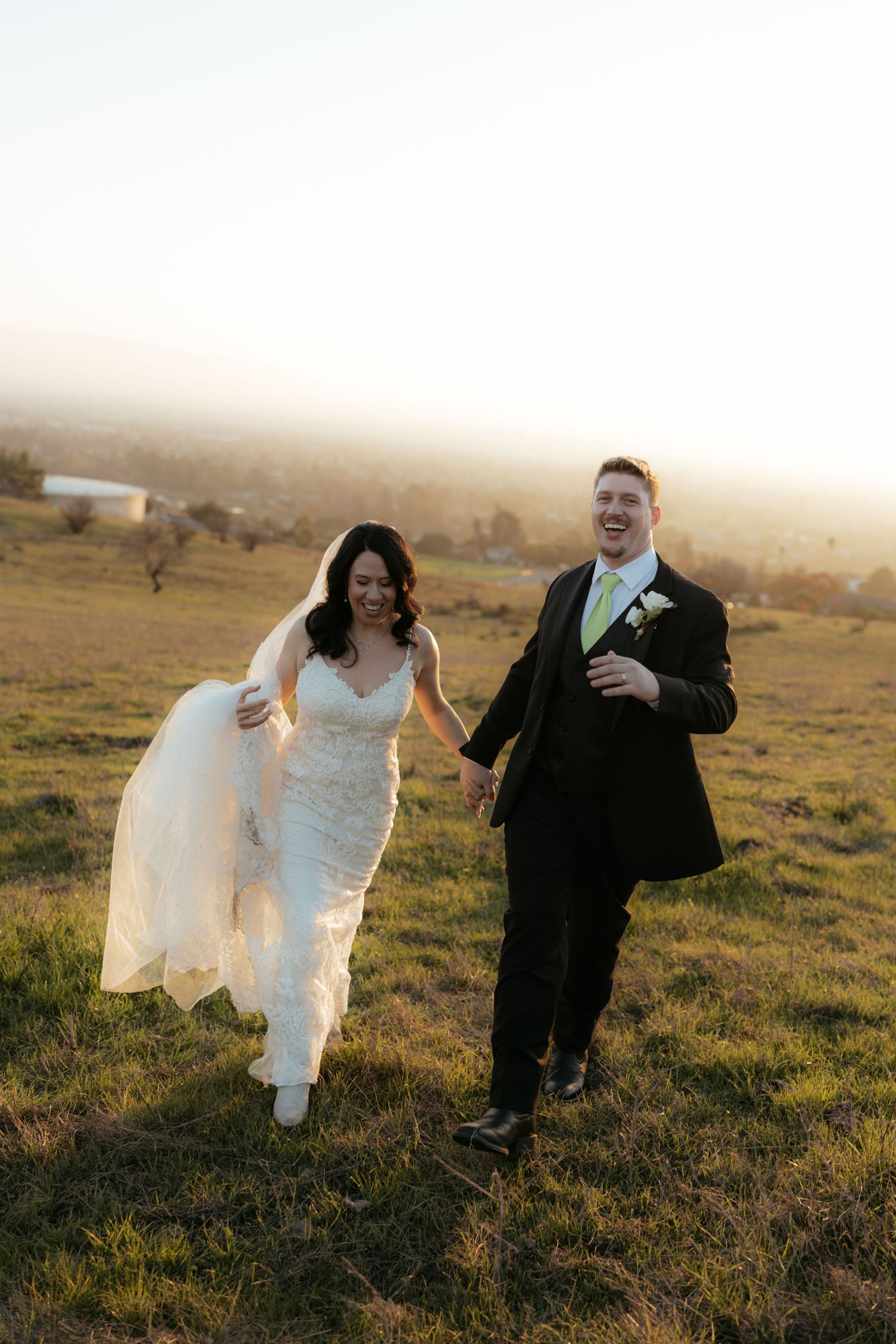 Playful newlyweds run and laugh together across a golden sunset field.