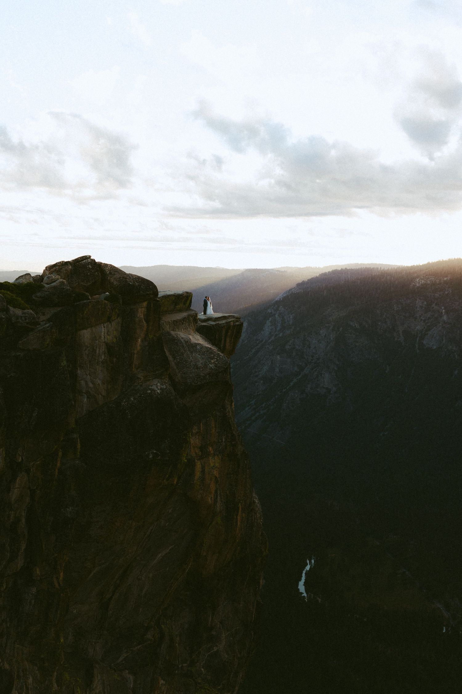 Two figures stand on a dramatic cliff edge at sunset, overlooking a vast mountain valley in Yosemite.