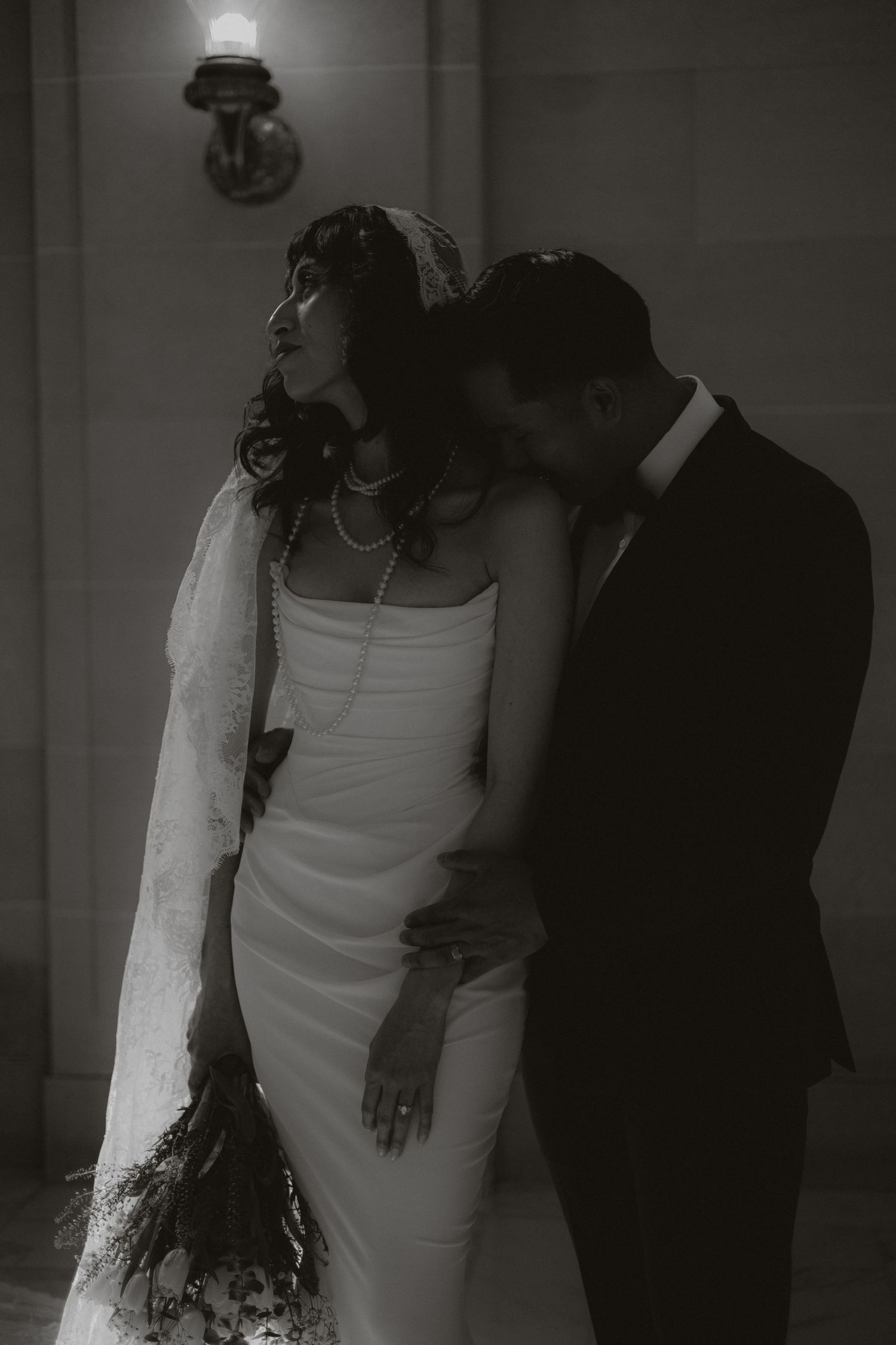 A moody black and white wedding photo of a couple in intimate embrace against a dimly lit wall.