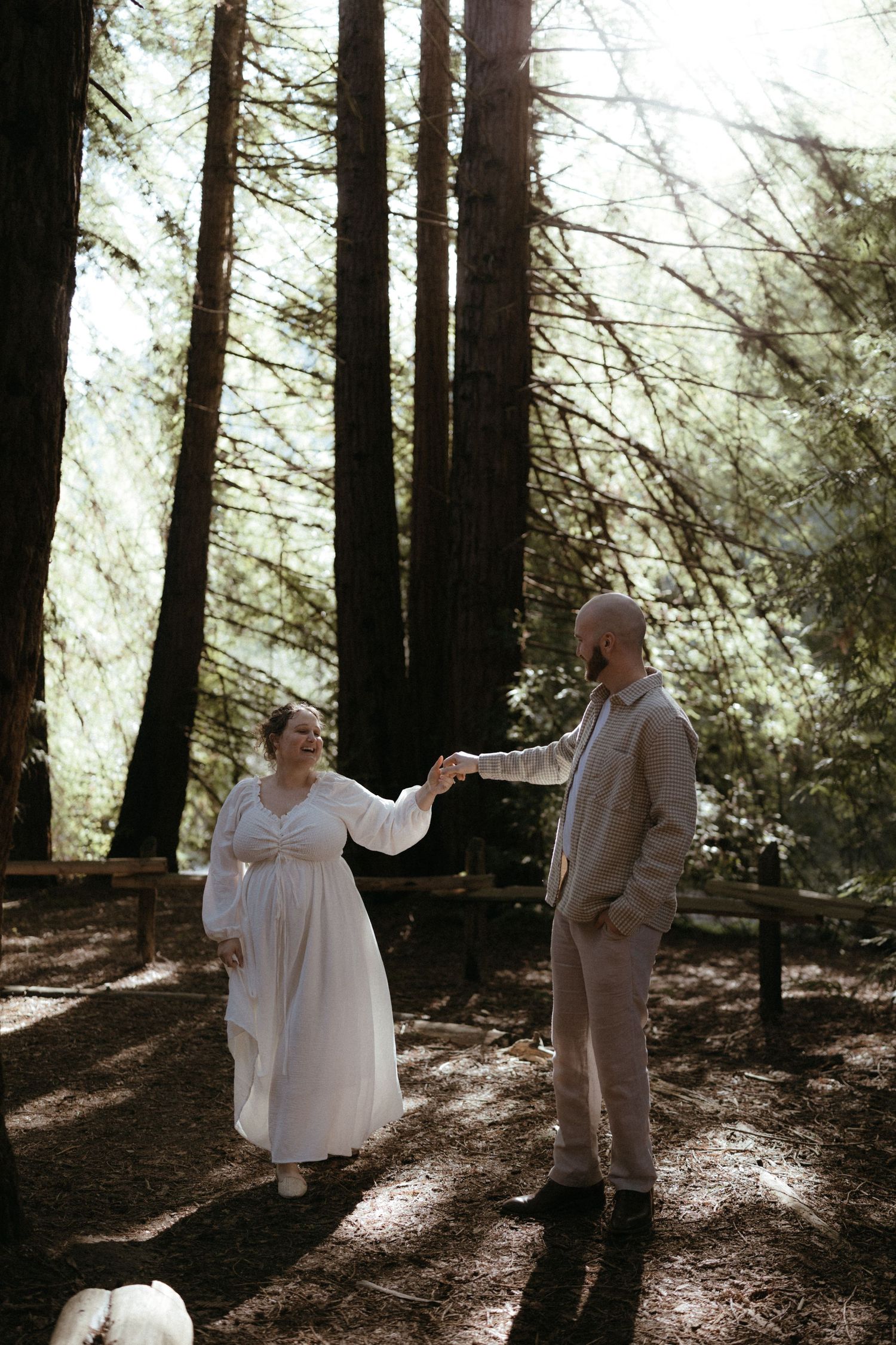 A couple shares an intimate dance in a sunlit redwood forest with rays of light streaming through the trees.