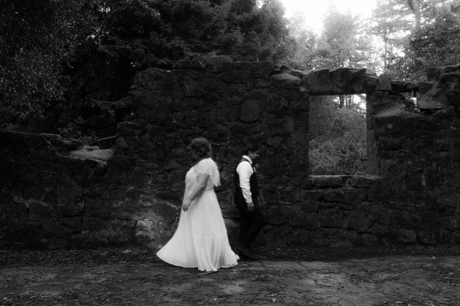 A black and white wedding photo series showing a couple walking through a garden path with stone walls.
